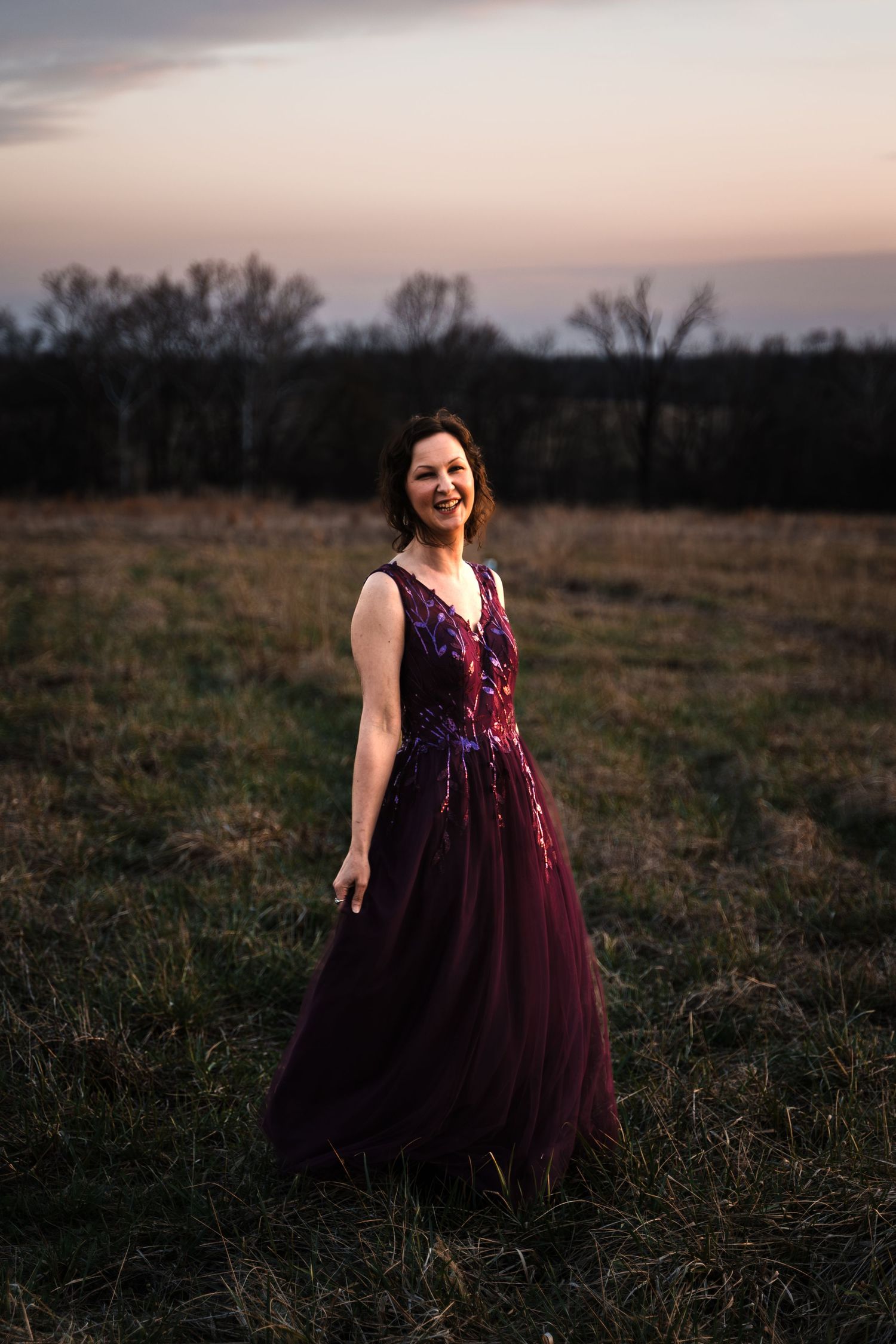 Bride smiling in stunning burgundy floral tulle gown standing in open field at dusk.