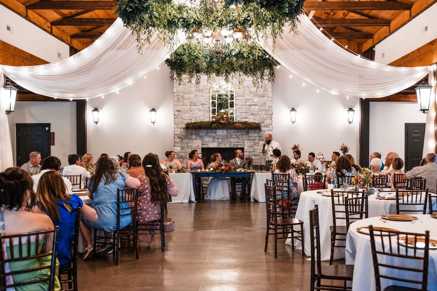 Elegant wedding reception in rustic barn with draped fabric ceiling, string lights, and stone fireplace.