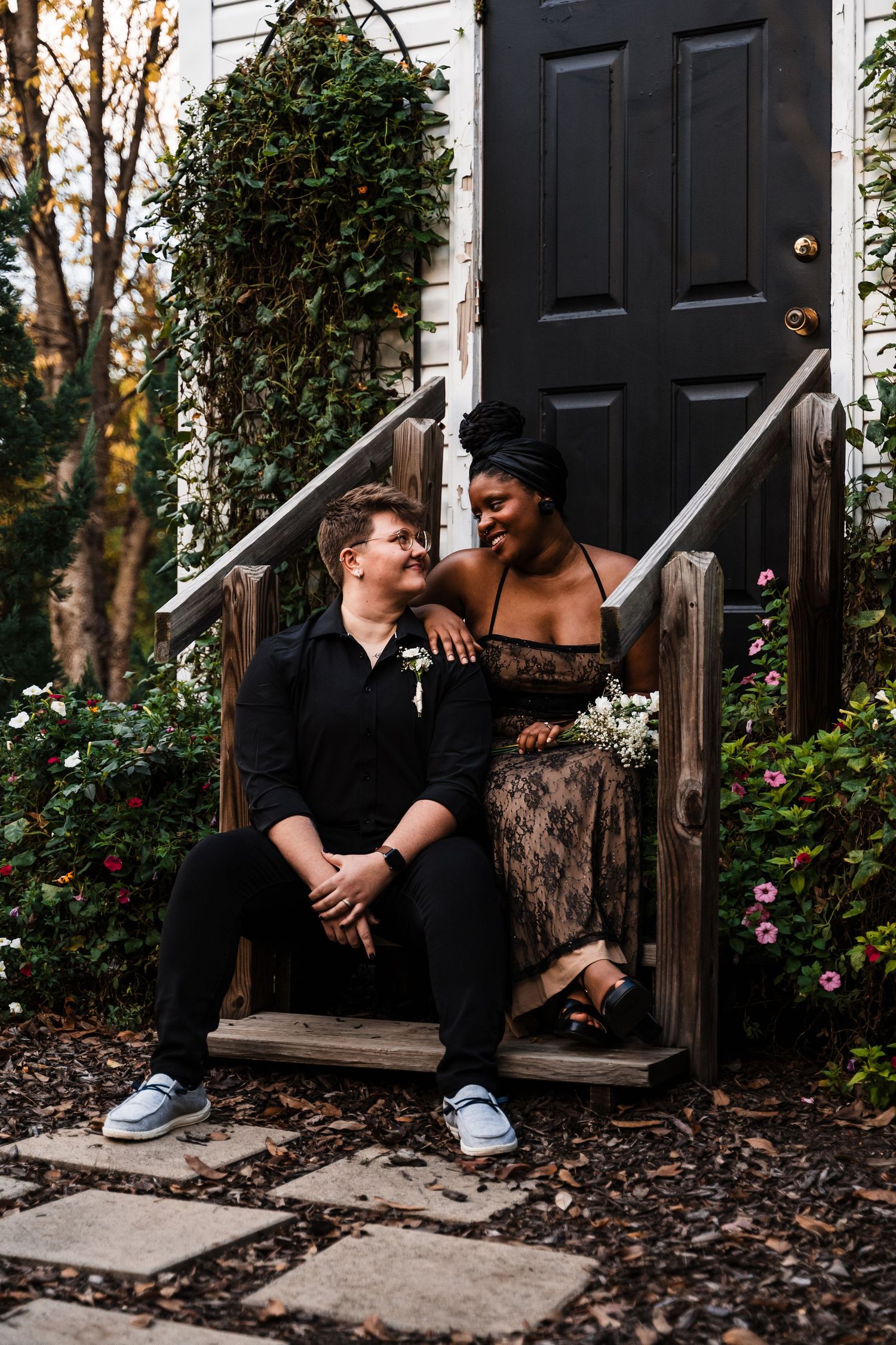 A couple sits intimately on outdoor steps by a black door with autumn leaves scattered around them.