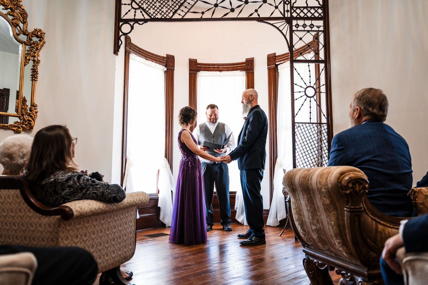 Couple exchanges vows before officiant in elegant Victorian room with ornate decor and seated wedding guests.