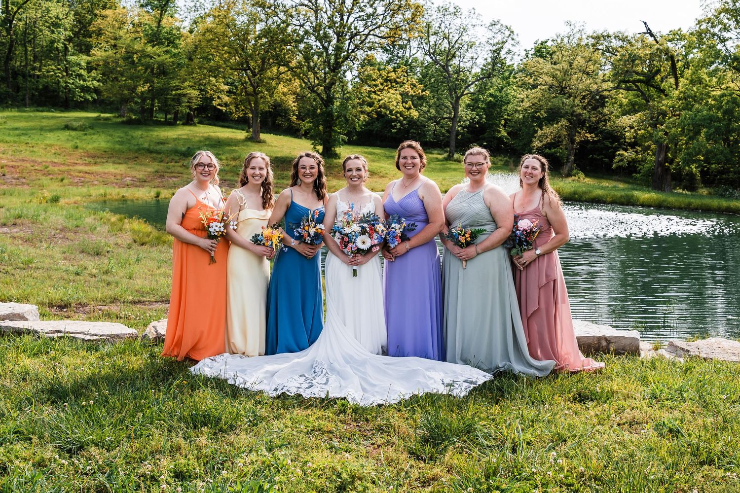 Bride in white gown poses with six bridesmaids in colorful mismatched dresses beside a scenic pond outdoors.