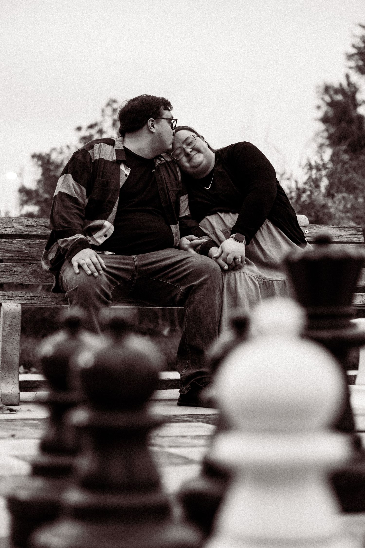A black and white artistic photo shows chess pieces in the foreground with two people resting on a bench in soft focus behind.