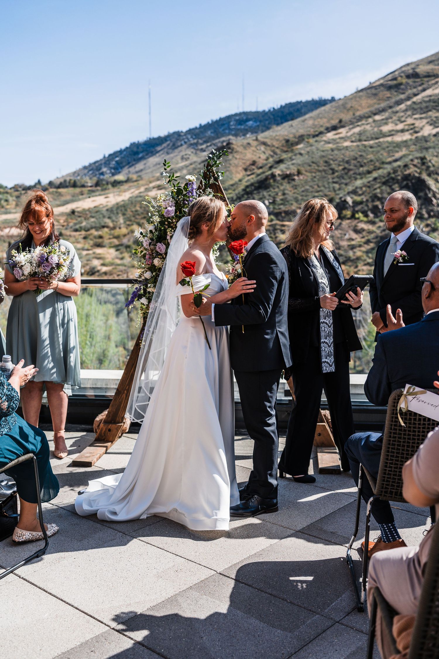 Bride and groom share first kiss at outdoor wedding ceremony with floral arch and mountain backdrop.