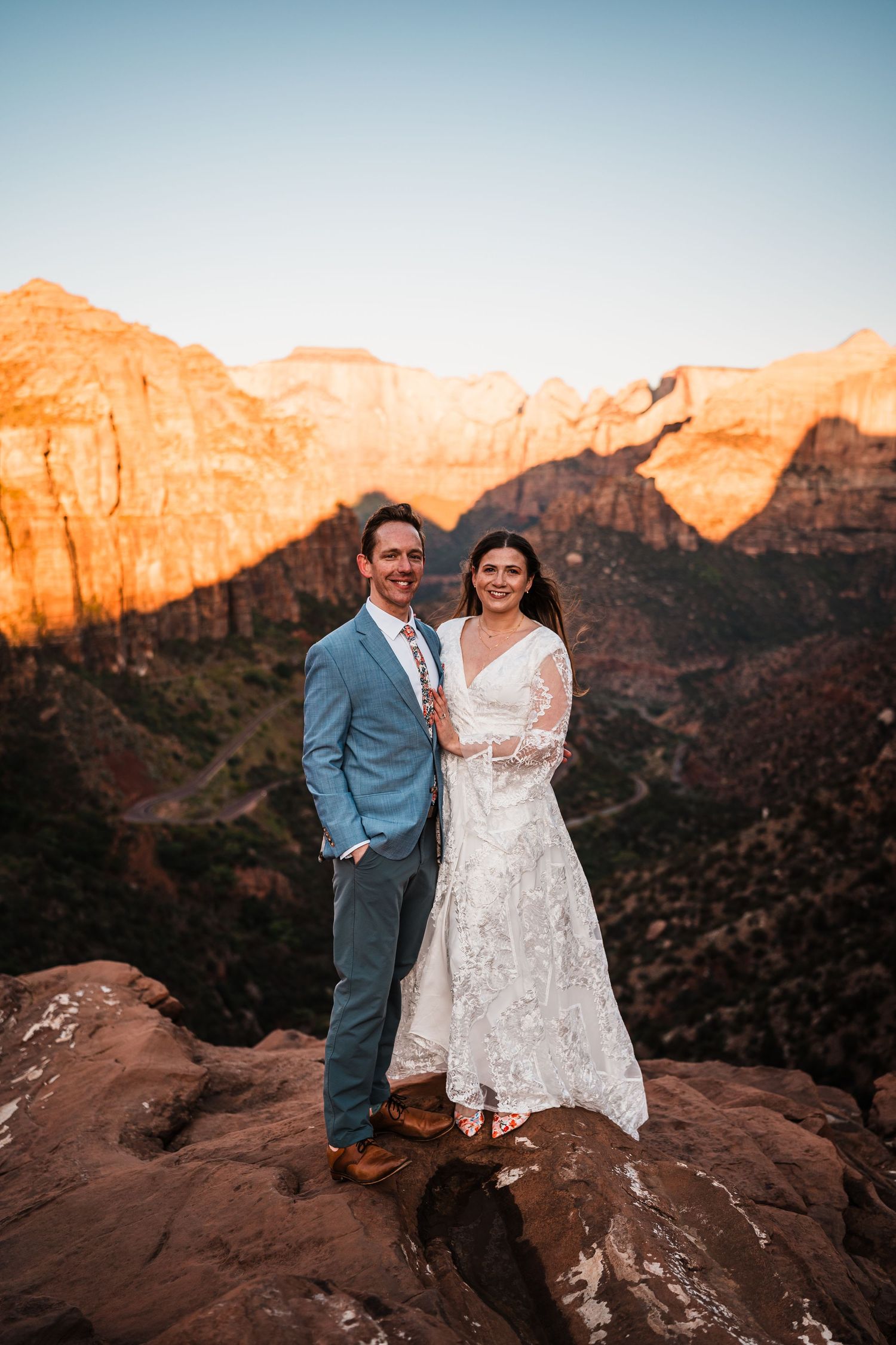 Smiling bride and groom posing on red rocks with stunning Zion National Park canyon backdrop at sunset.