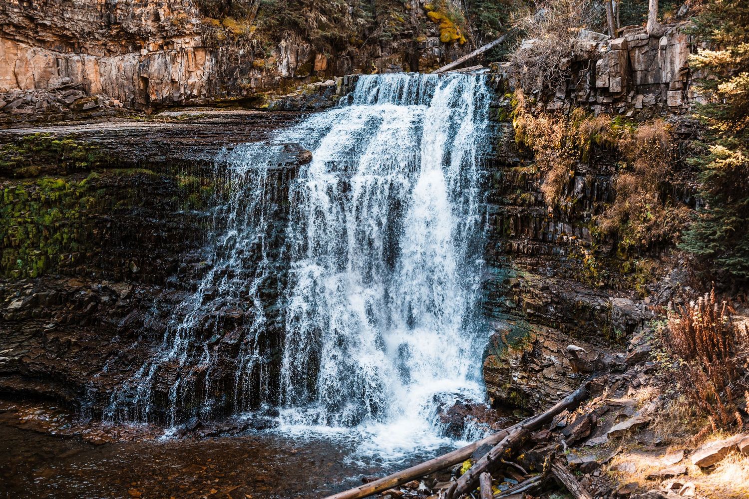 A cascading waterfall flows down rocky cliffs surrounded by autumn foliage in a natural landscape.