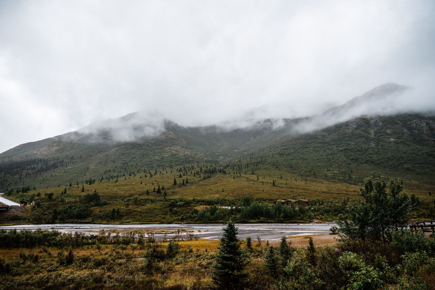 Misty mountains rise above a serene lake surrounded by autumn-colored vegetation in a moody landscape scene.