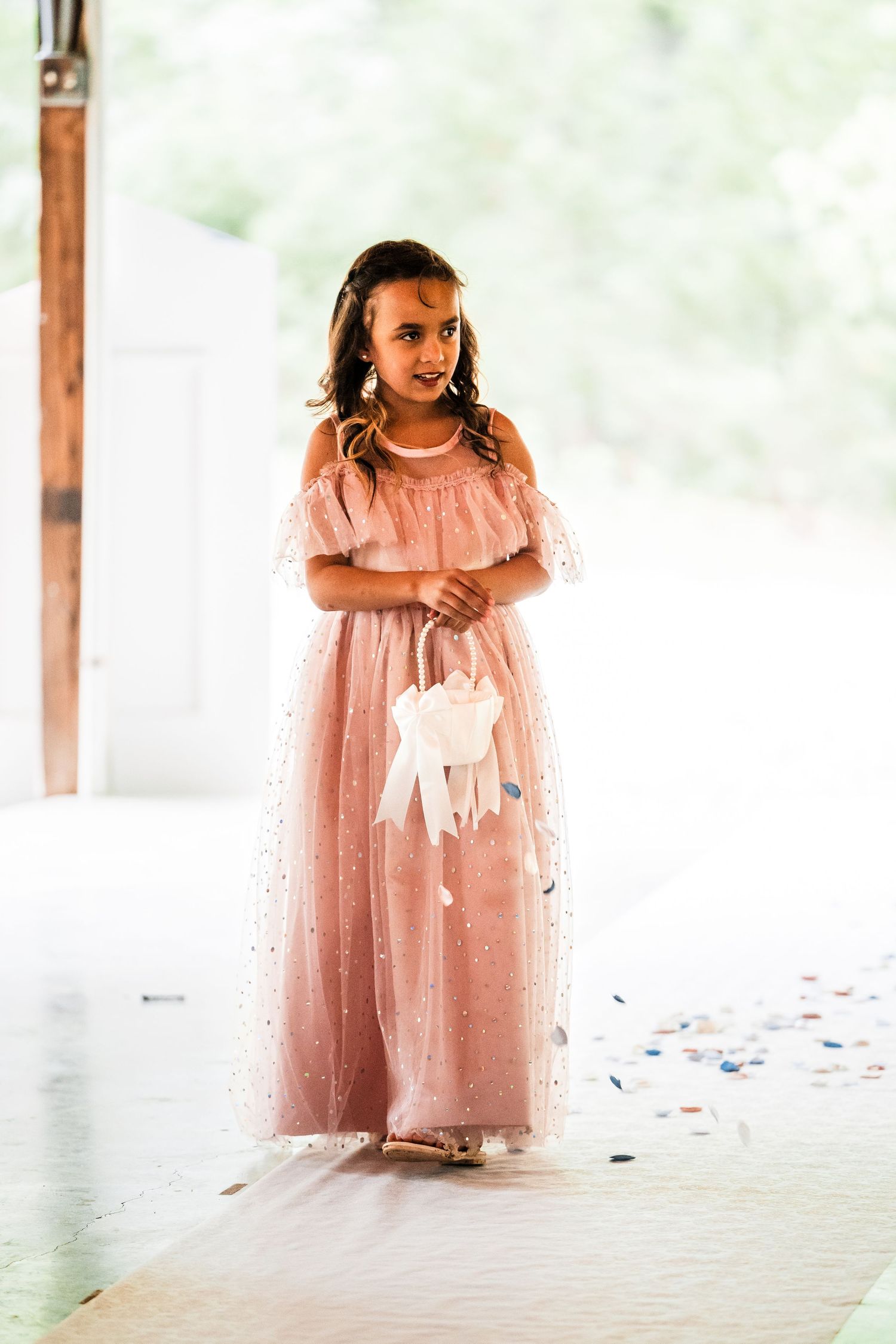 Young attendant in a flowing peach dress holds white object during wedding ceremony.