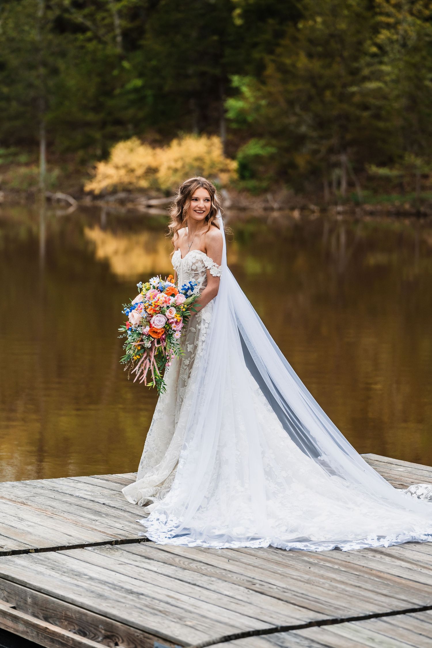 Stunning bridal portrait on a wooden dock with colorful bouquet and long train reflecting in lake water.