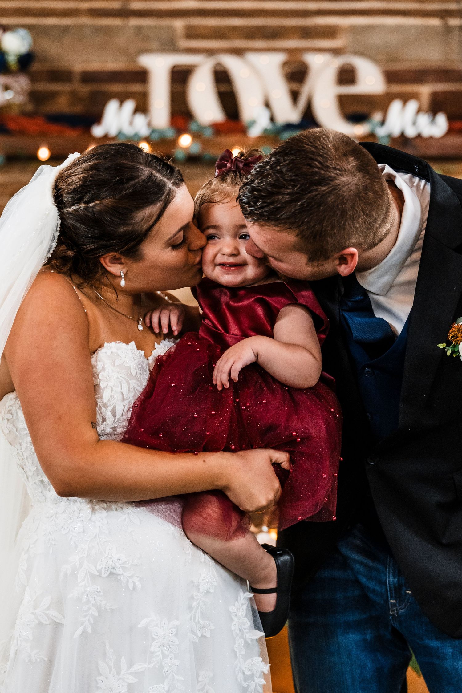 Sweet candid moments between wedding couple and child in burgundy dress at reception.