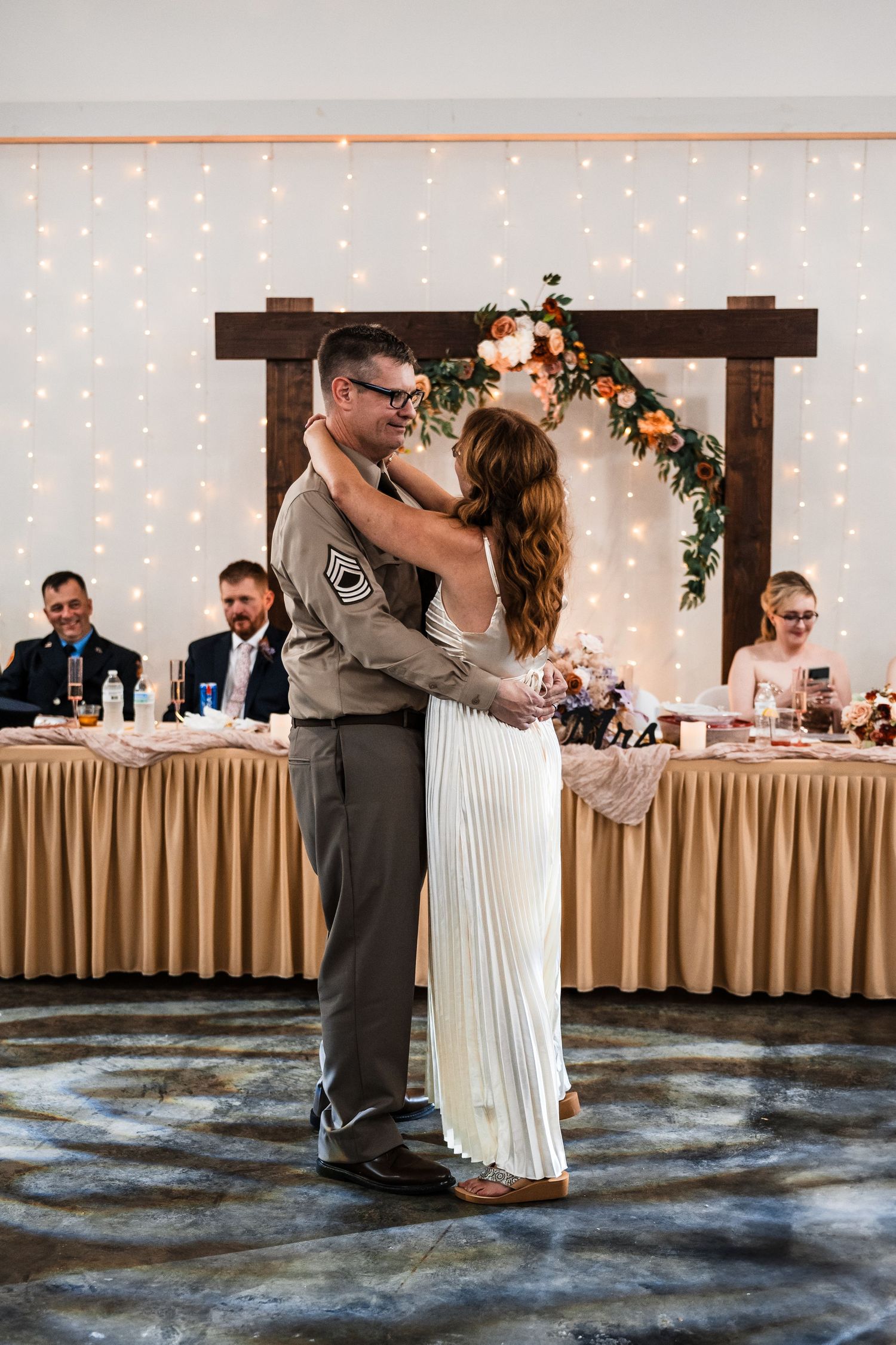 A couple shares their first dance at a wedding reception with string lights and floral arch decorations.