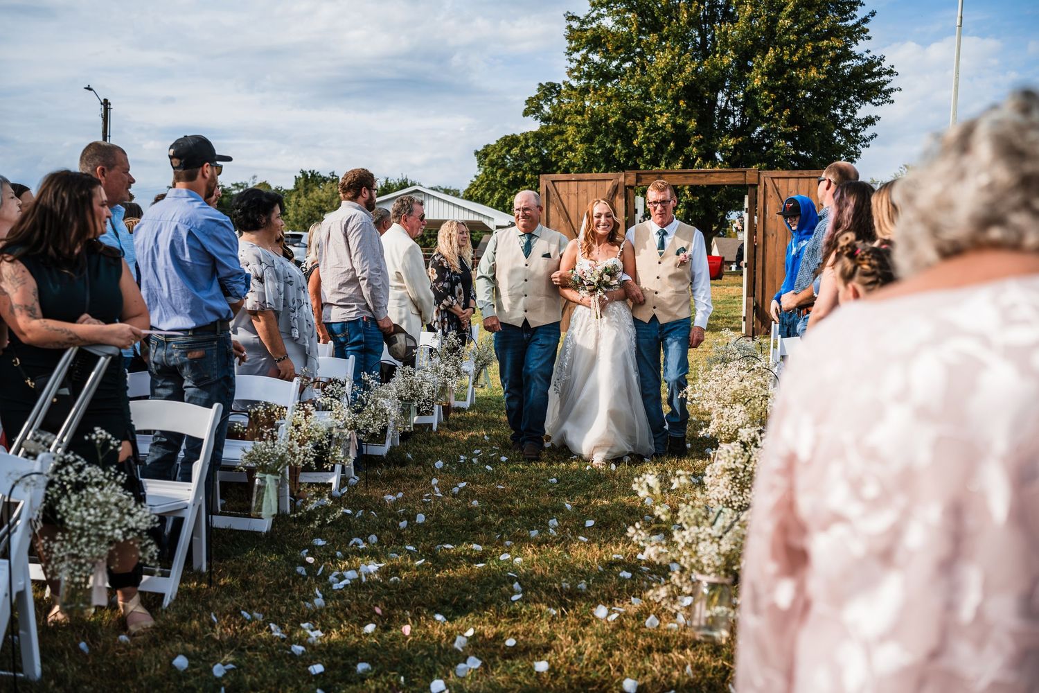 Guests watch an outdoor wedding ceremony with white chairs and floral decorations.