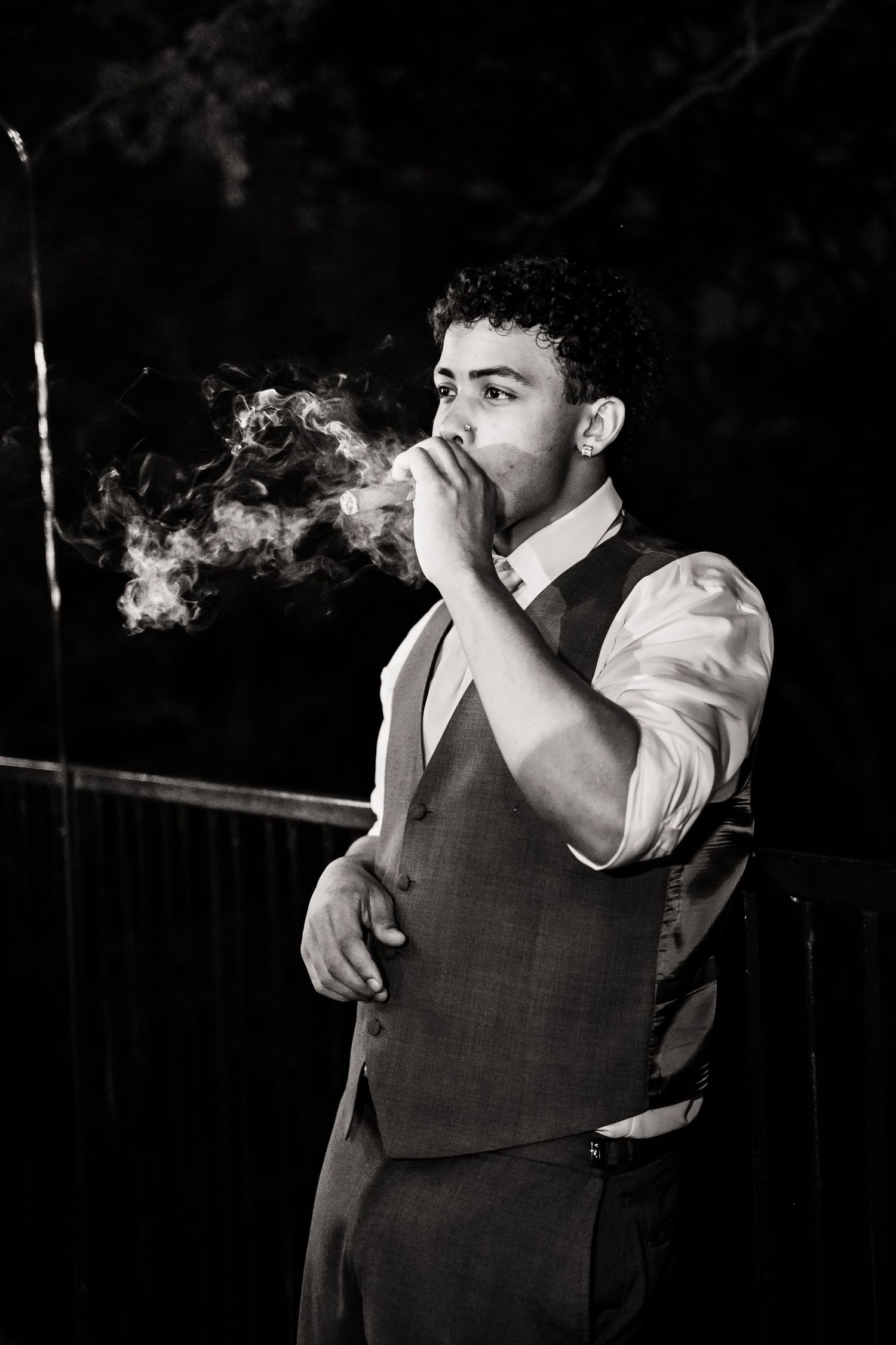 A black and white portrait of someone in a vest and dress shirt exhaling cigarette smoke in dramatic lighting.
