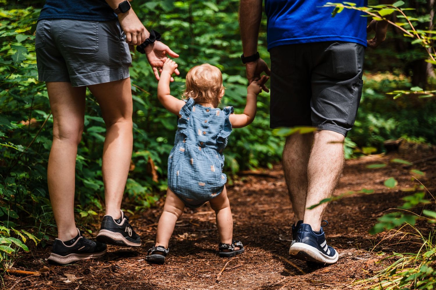 Two adults helping a small child walk along a forest trail.