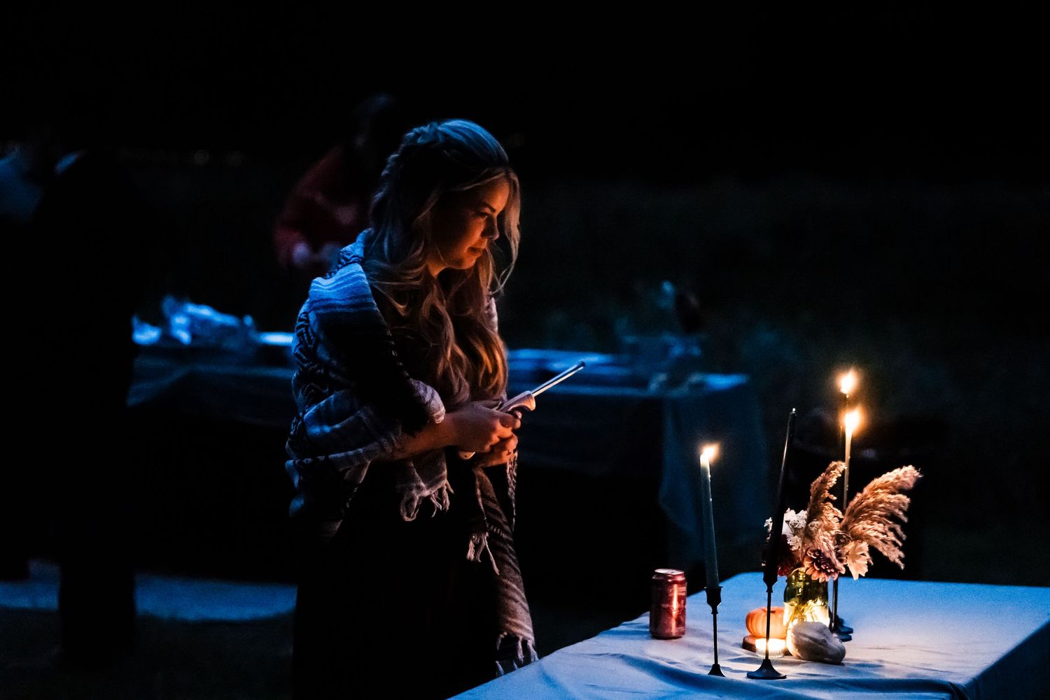 A moody candlelit scene shows a person gazing down at table decorations in dark atmospheric lighting.