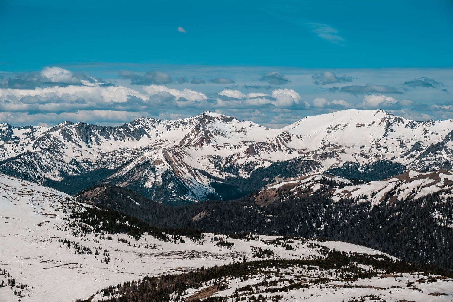 Snow-covered Rocky Mountain peaks stretch across the horizon under a bright blue sky with scattered clouds.