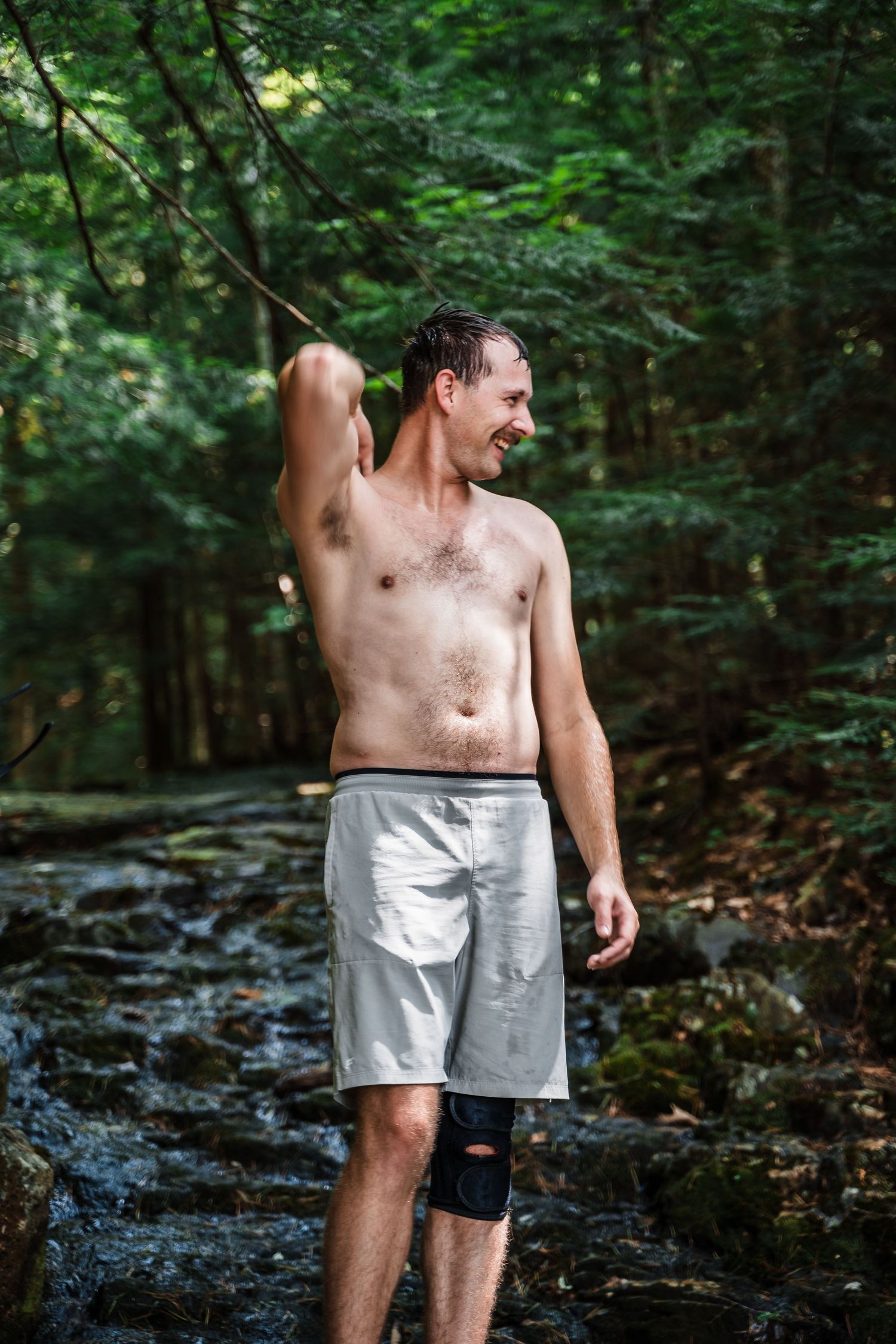 Shirtless hiker with knee brace stands in a rocky forest stream on a sunny day.