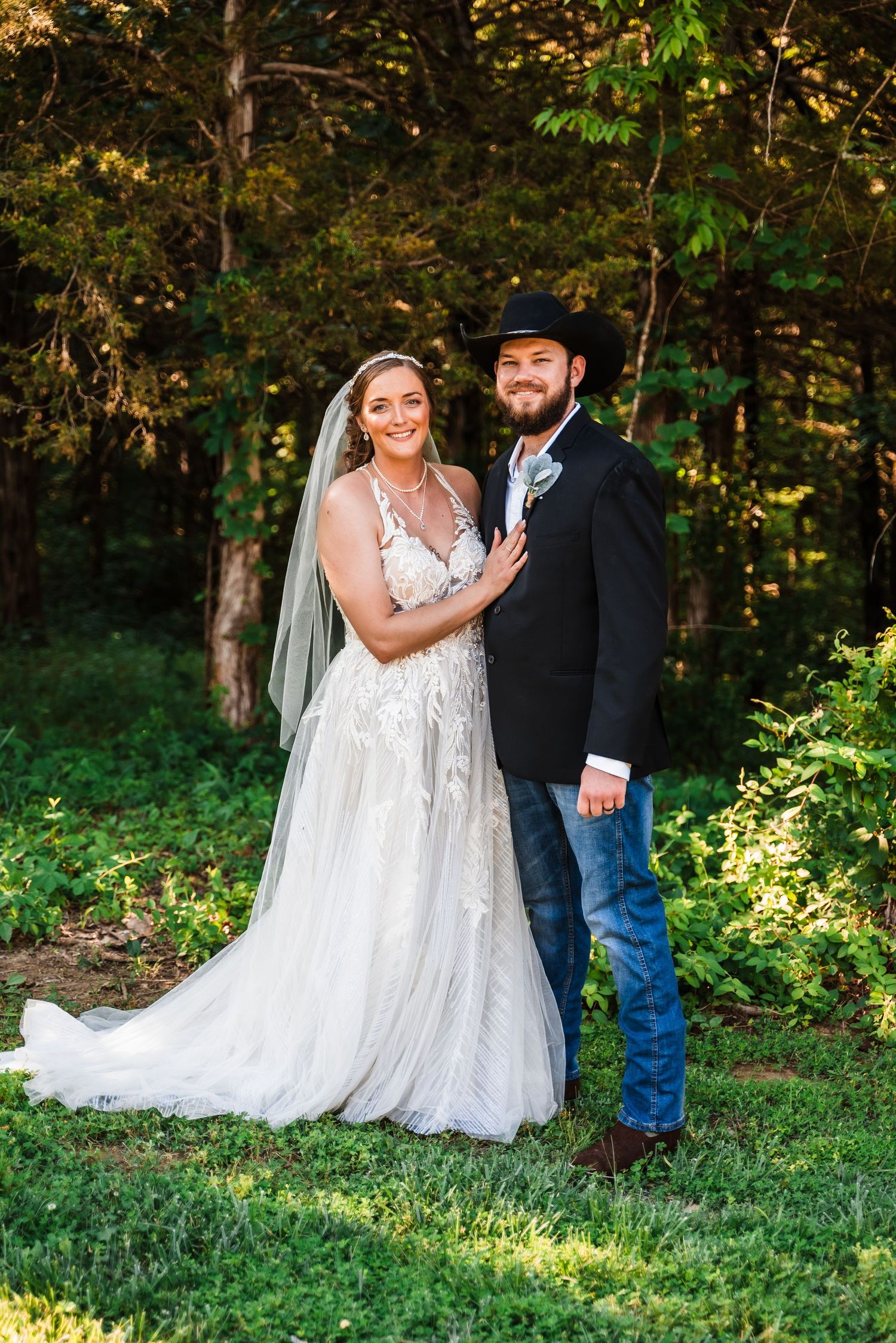 A couple poses for a wedding photo outdoors wearing a white gown and black jacket with cowboy hat in a wooded setting.
