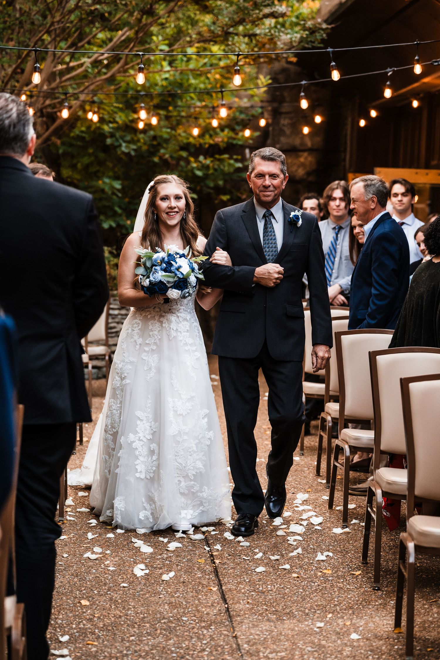 Ceremony processional under string lights with white flower petals lining the aisle.
