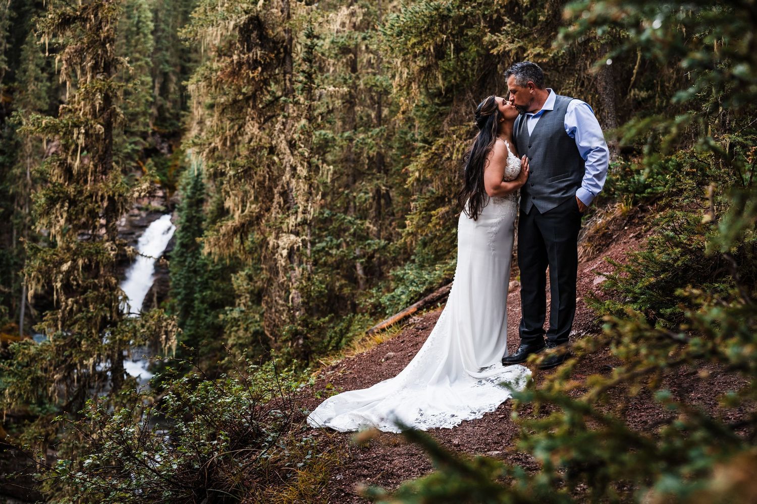 Bride and groom sharing a kiss on a forest trail with a waterfall visible in the background.