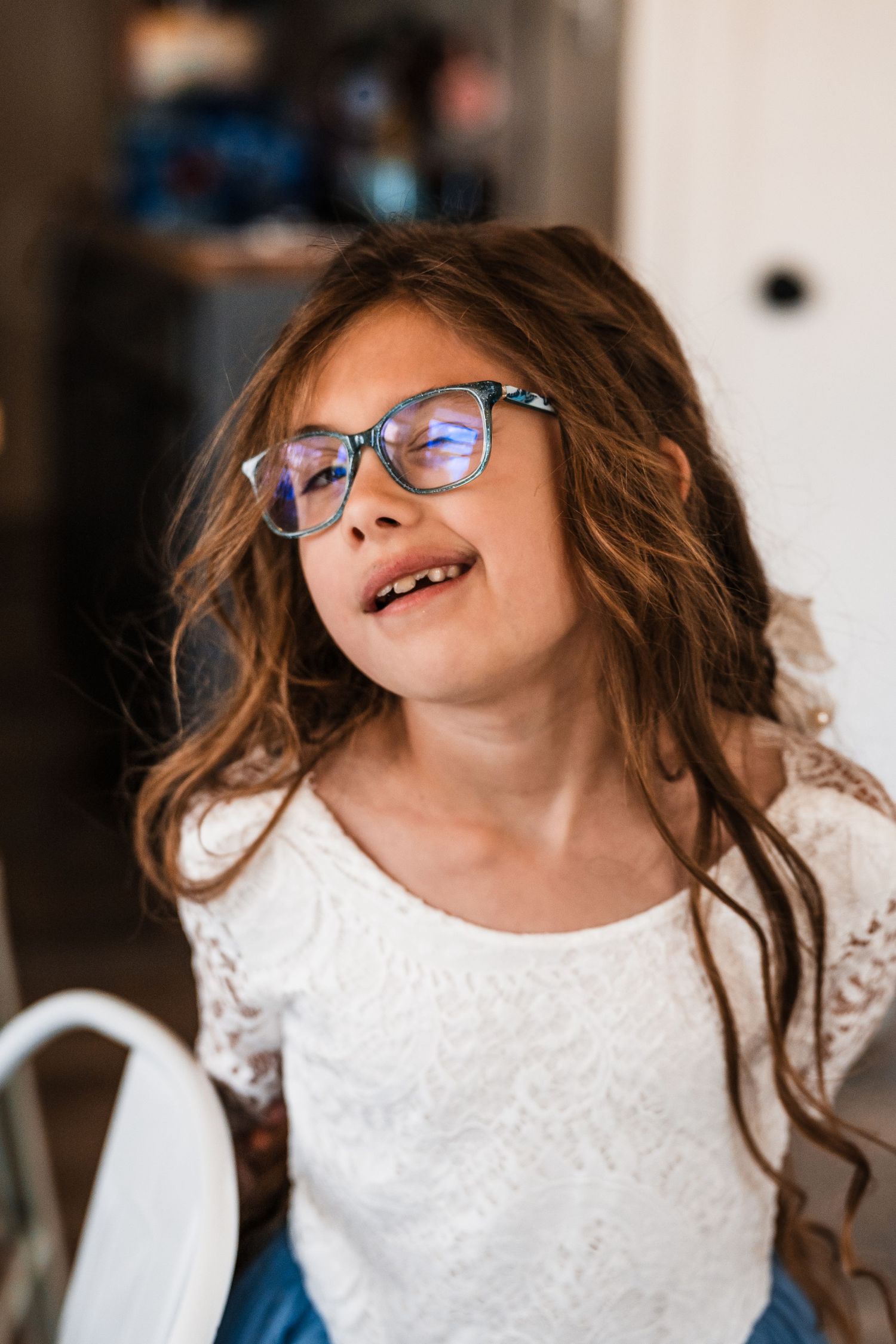 Young girl with long brown hair and blue glasses wearing a white lace top, making a funny face indoors.