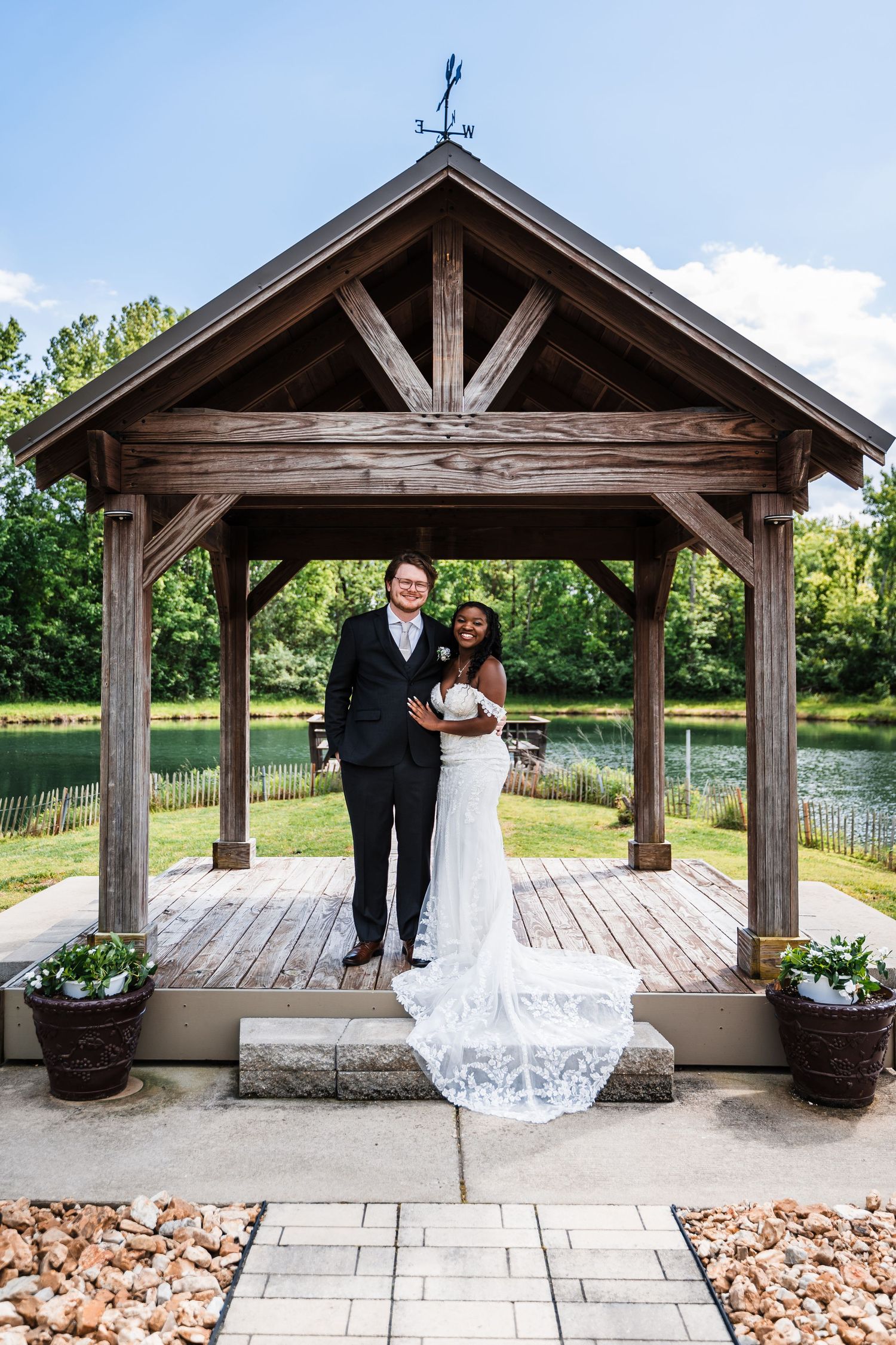 Interracial wedding couple posing under a rustic wooden gazebo beside a serene pond outdoors.