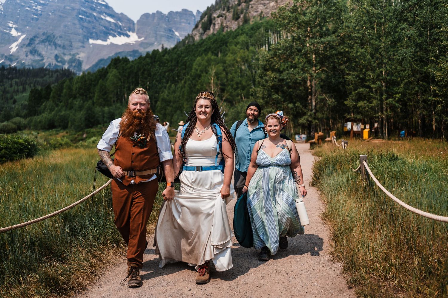 Wedding party walks path near Maroon Bells mountains, bride in white dress with groom in brown vest.