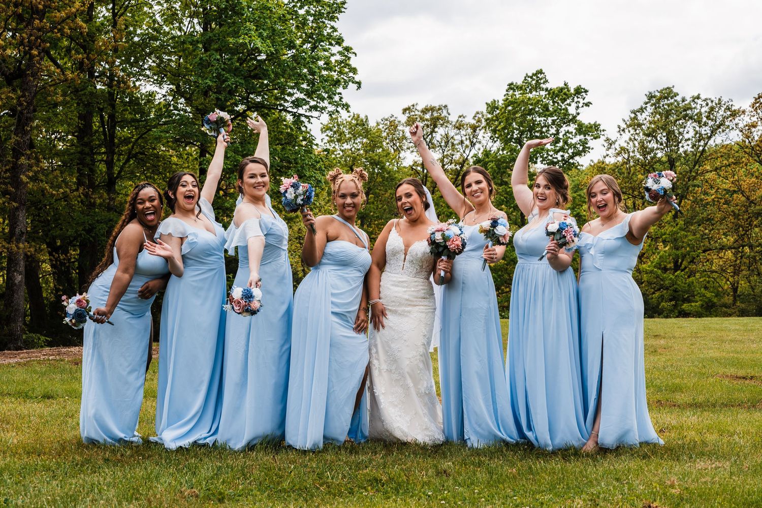Bride and bridesmaids in light blue dresses celebrating with raised arms on a sunny day.