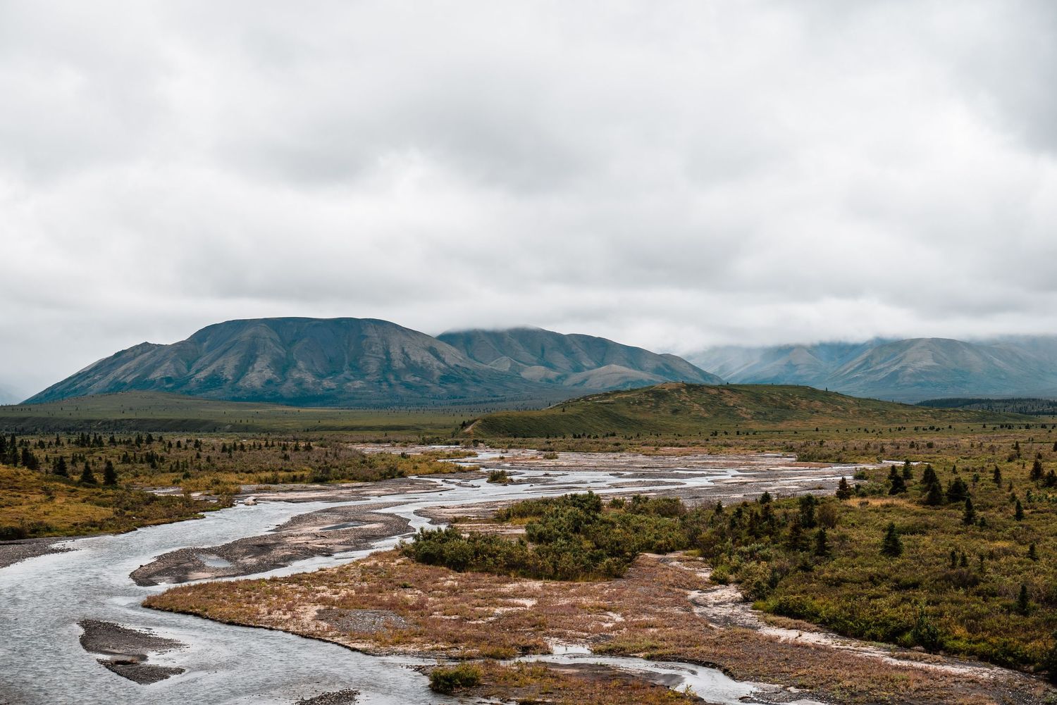 A winding river flows through rugged mountains and tundra landscape under cloudy skies in Alaska.