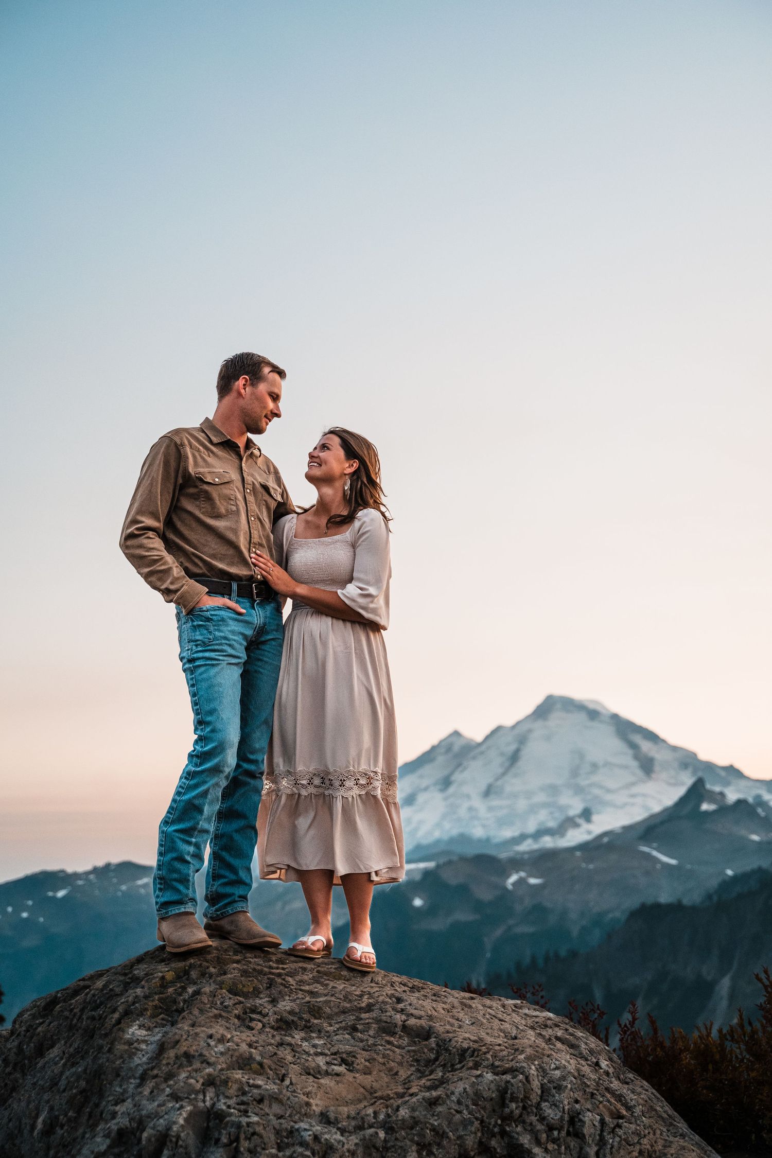 A couple stands together on a rocky overlook during sunset with a snow-capped mountain peak in the distance.