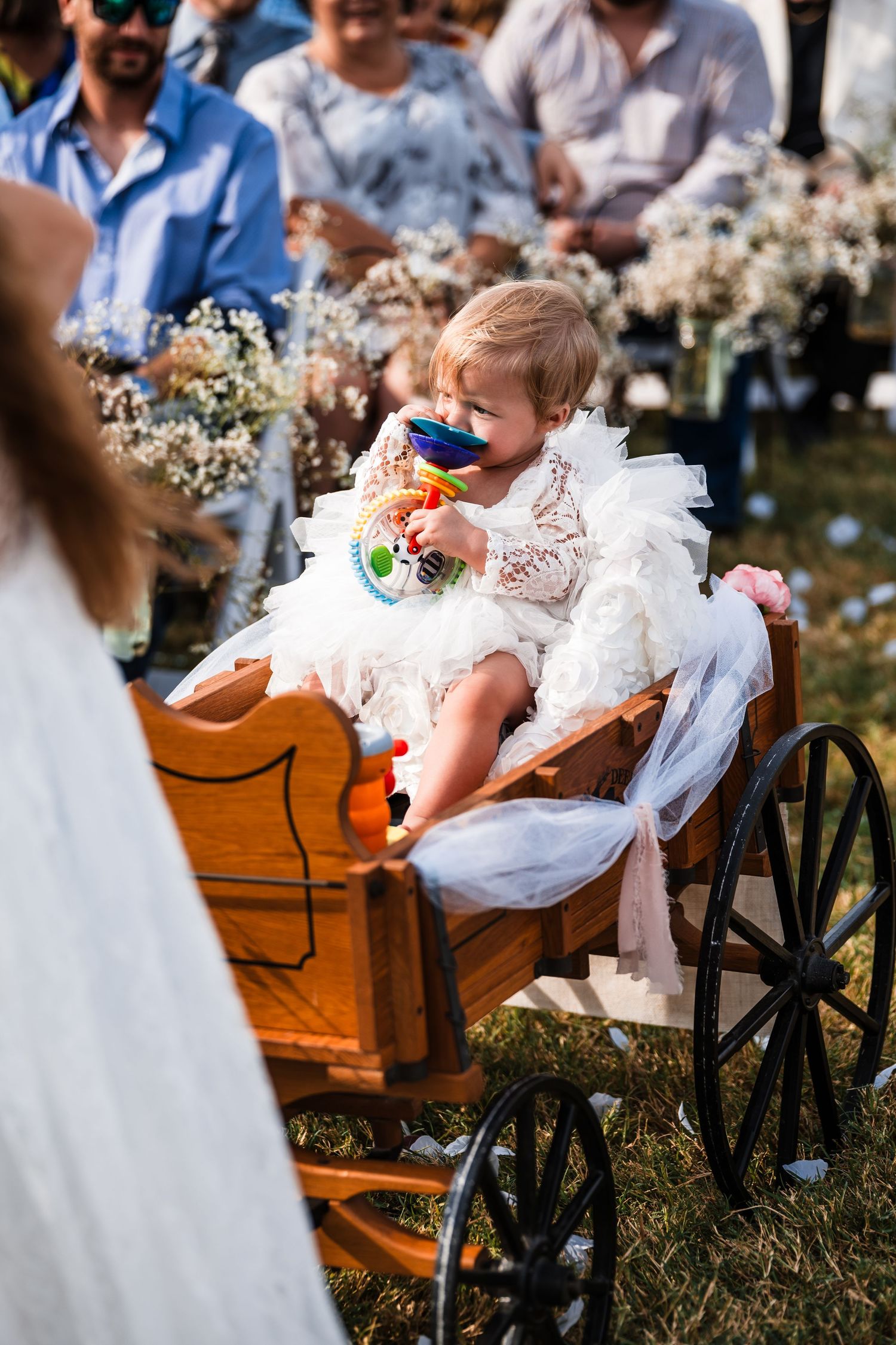 A child in a white dress sitting in a rustic wooden wagon during an outdoor wedding.