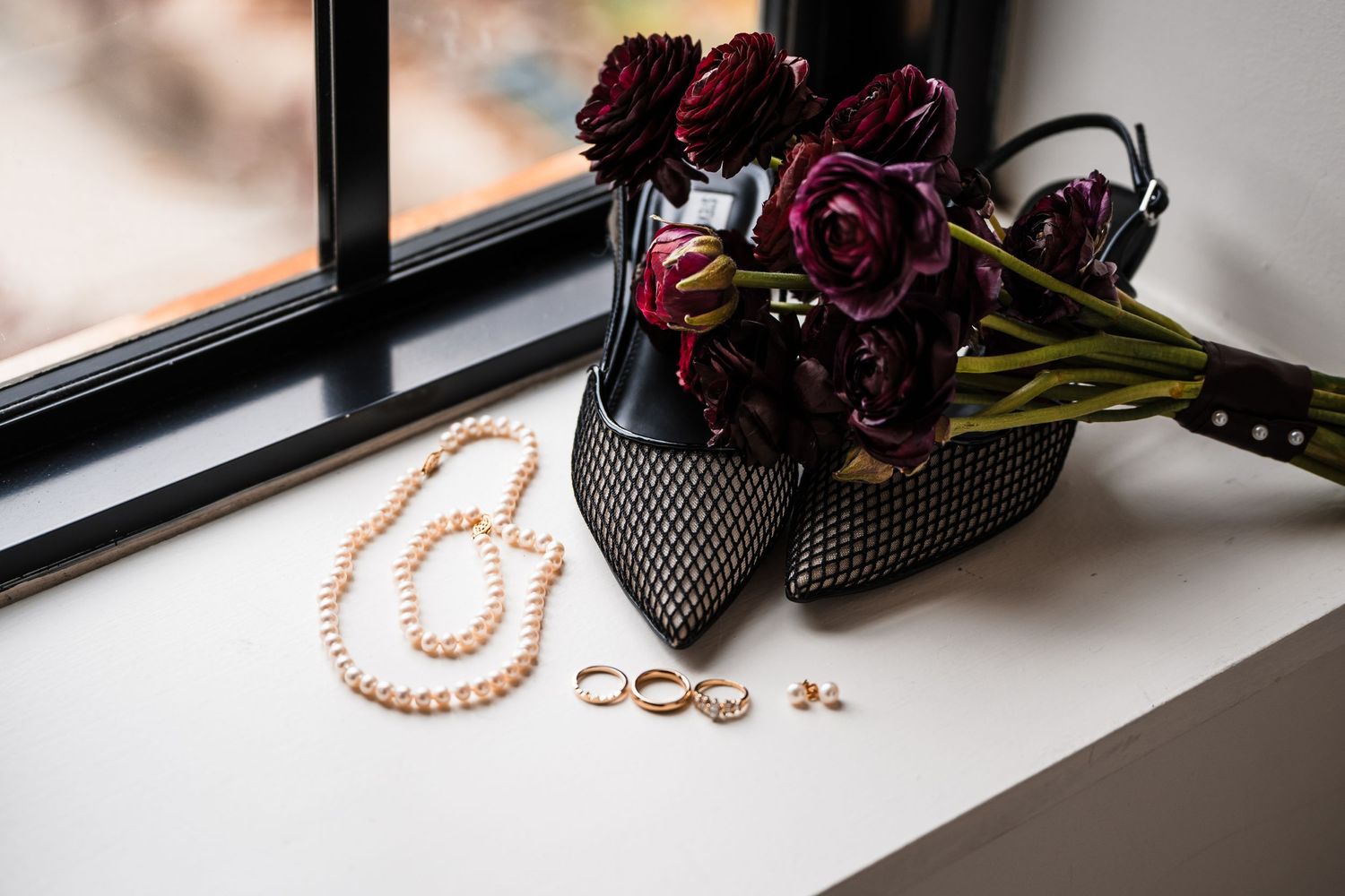 Wedding accessories and dark purple flowers arranged on a windowsill with rings and pearl necklace.