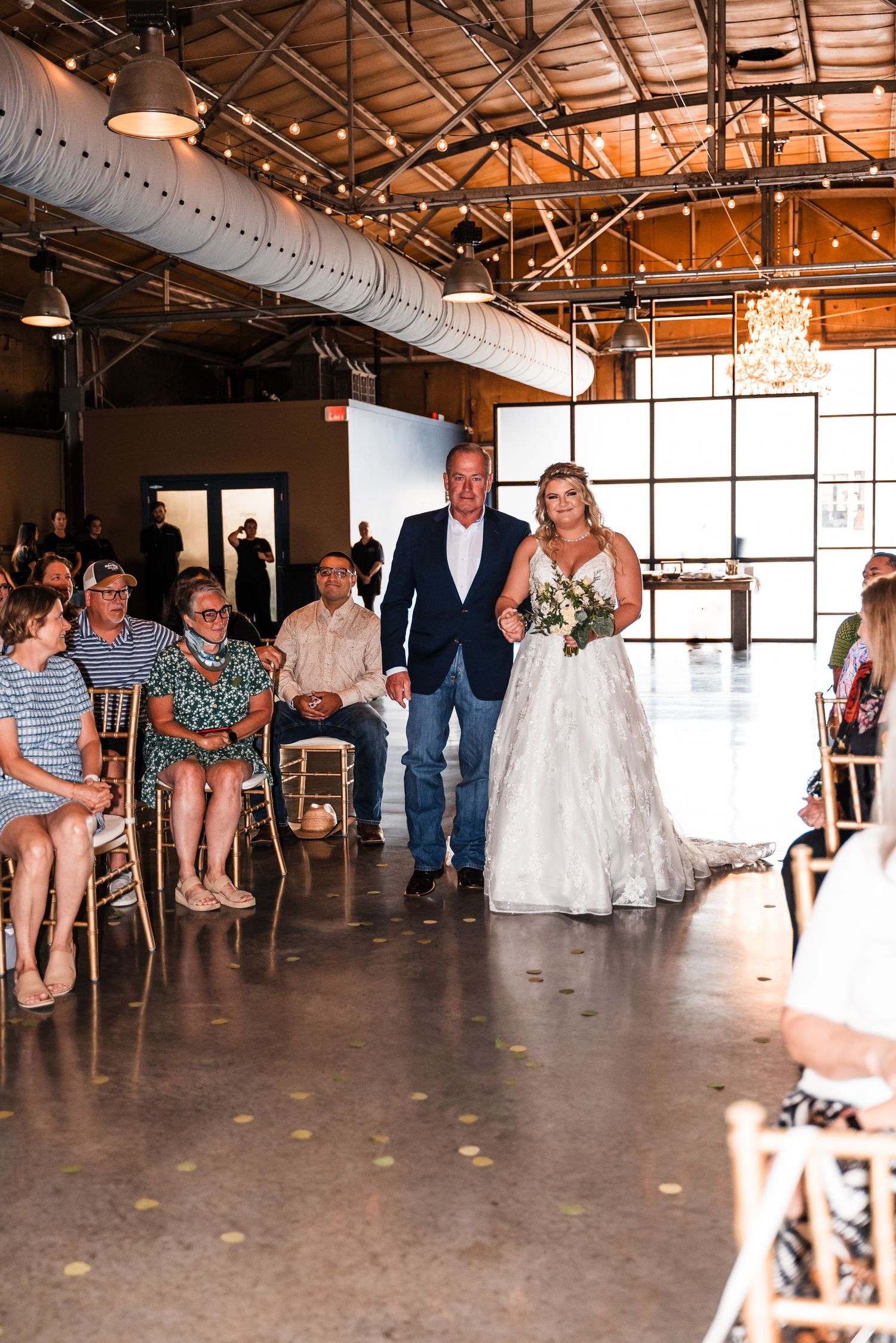 A wedding celebration takes place in an industrial-style venue with exposed wooden beams and large windows.