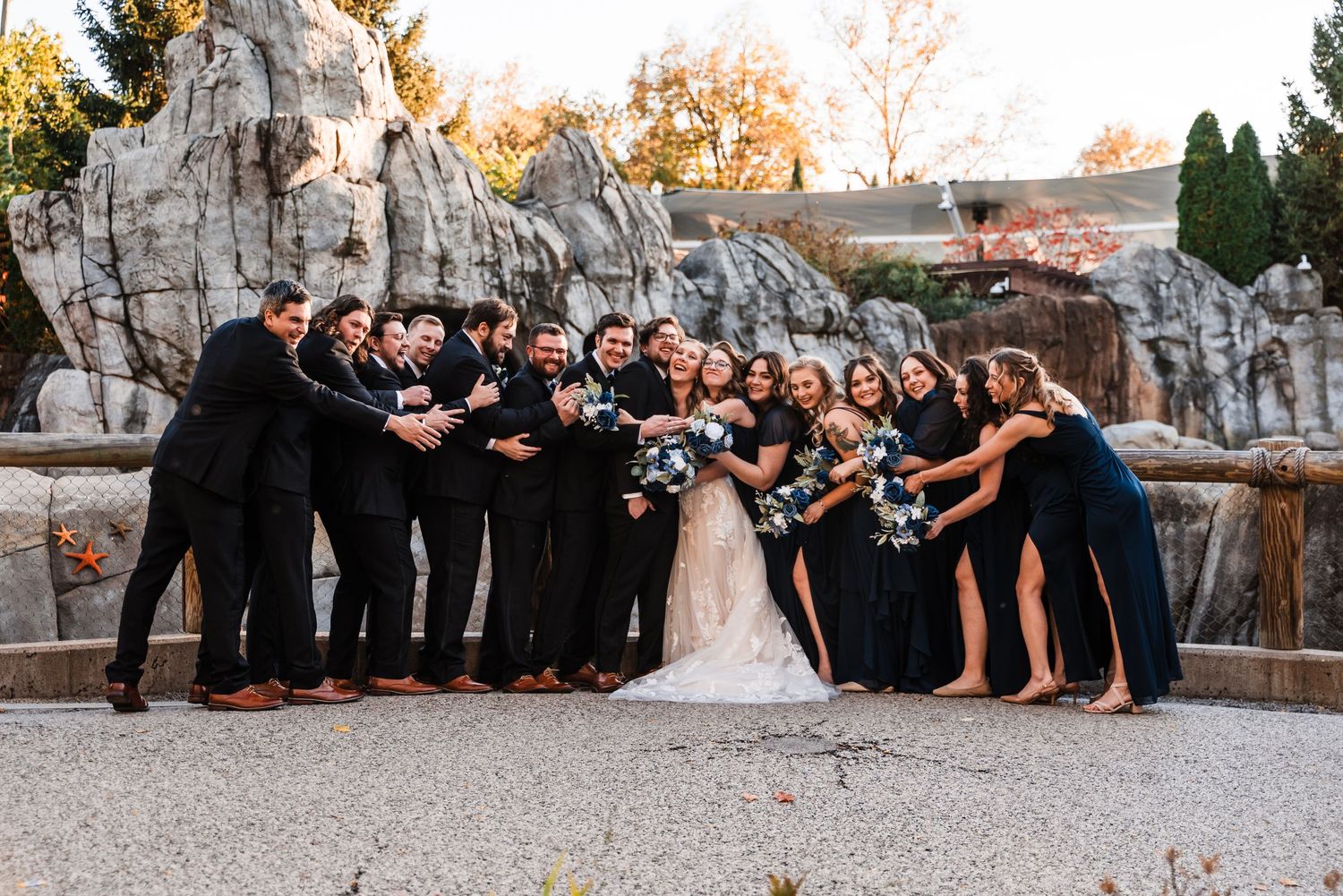 Large wedding party poses together in formal black attire against stone backdrop.