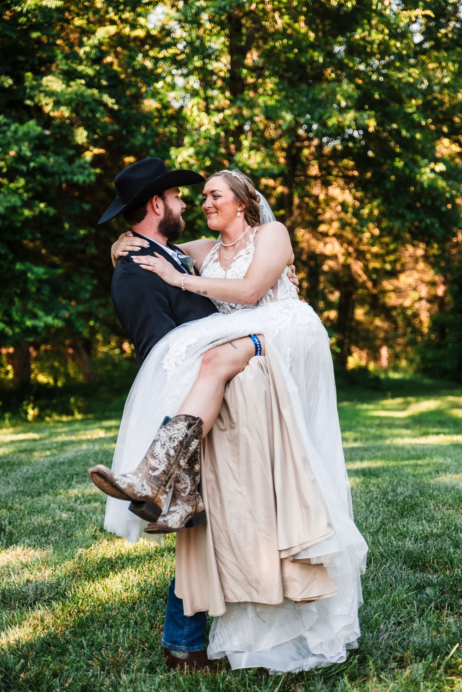 A joyful wedding couple in a sunlit garden, with the bride wearing cowboy boots under her flowing white dress.