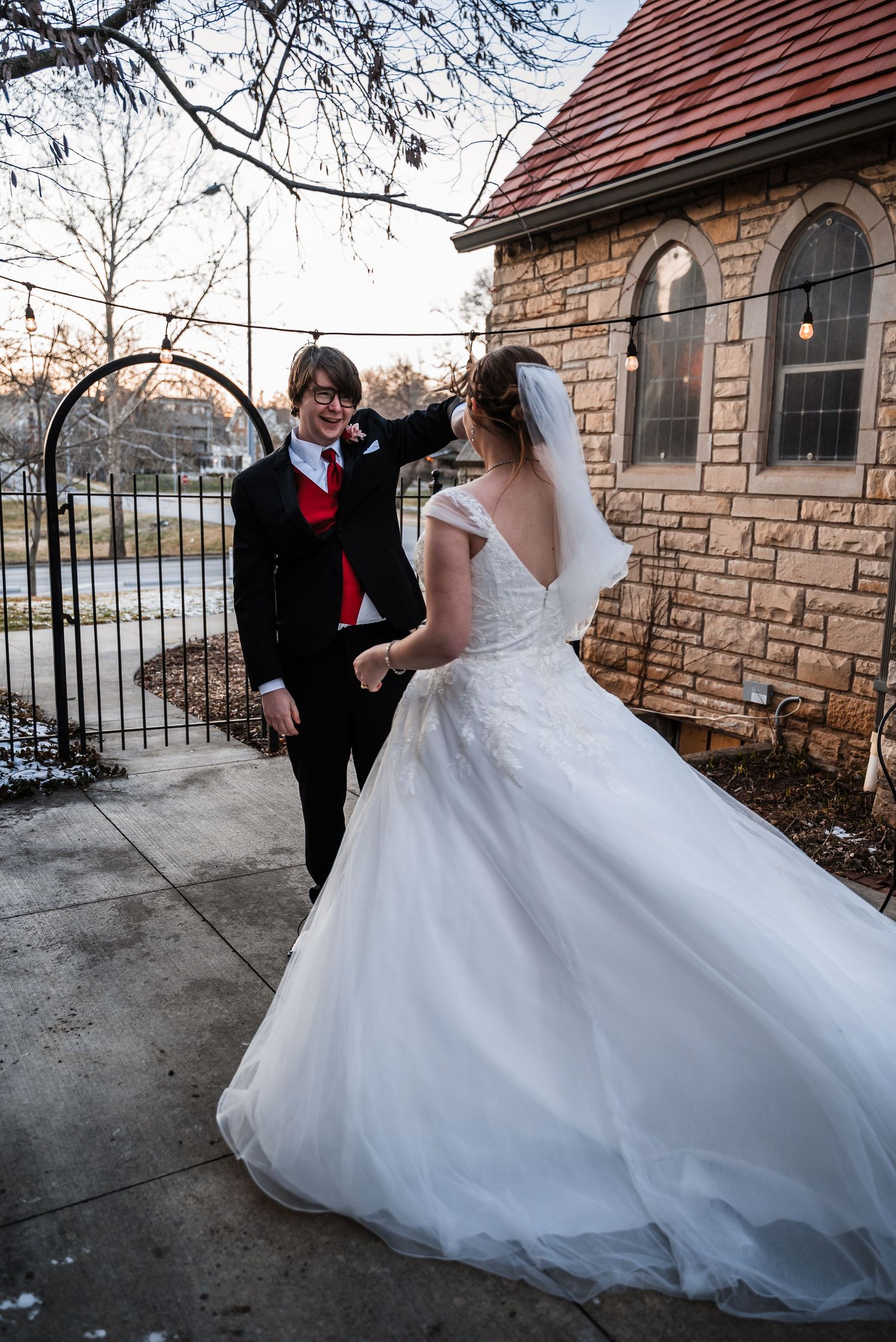 A joyful wedding couple dances outside a stone chapel with bare winter trees in the background.