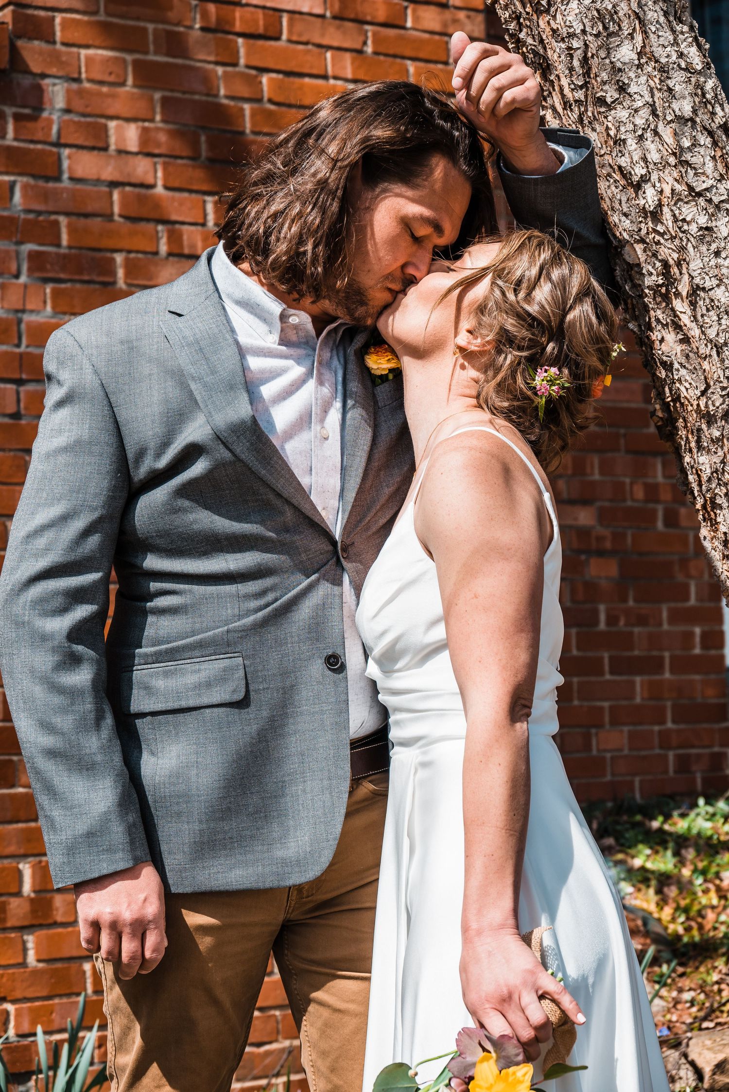 Romantic wedding couple embracing against brick wall on sunny day.