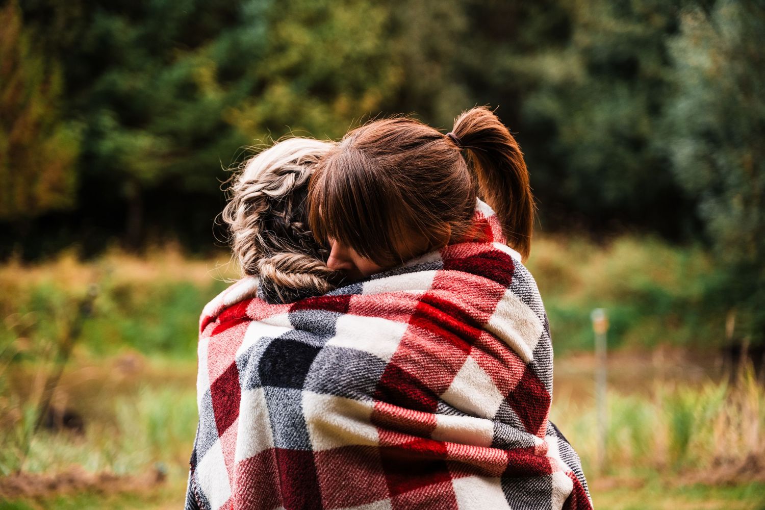Two figures wrapped in a cozy plaid blanket outdoors on an autumn day.