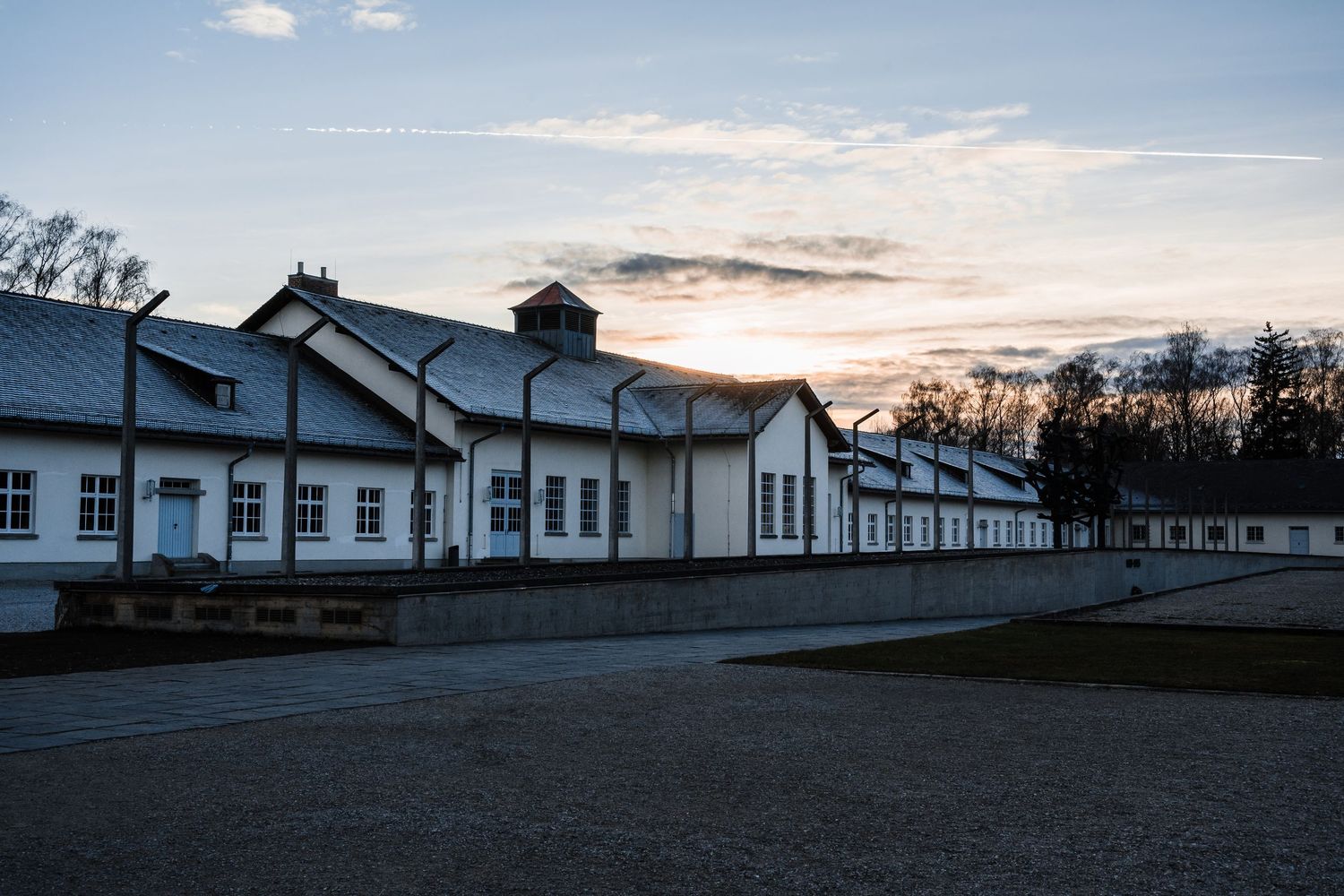 White barracks buildings at dusk with silhouetted trees against a fading sunset sky at a historical site.