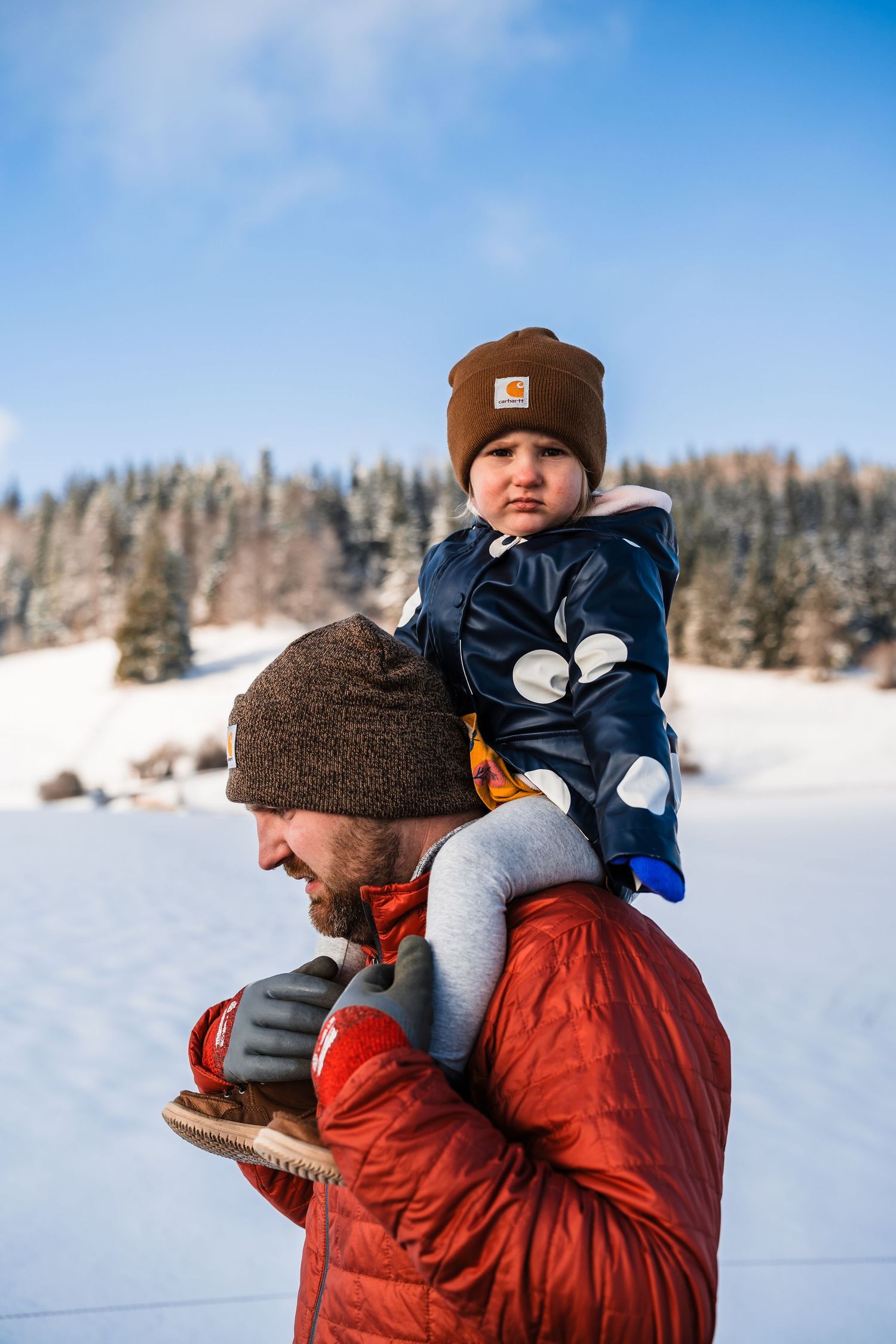 A parent carries a child wearing a brown beanie and navy coat on their shoulders in a snowy winter landscape.