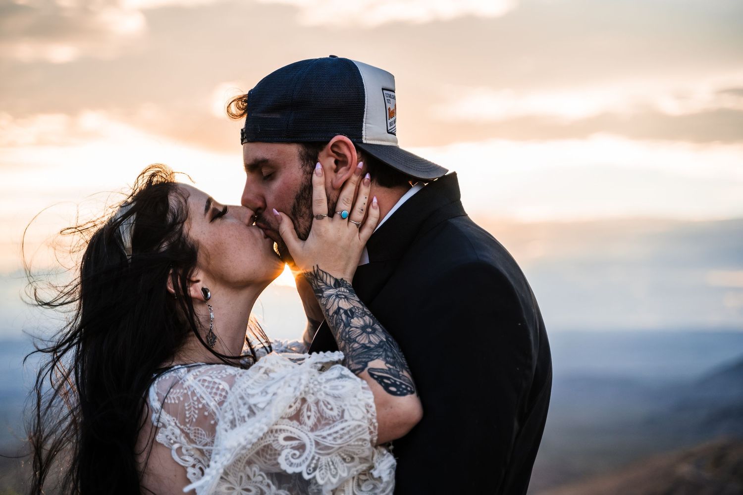A couple shares a romantic kiss on a mountain summit during sunset with dramatic cloudy skies in the background.