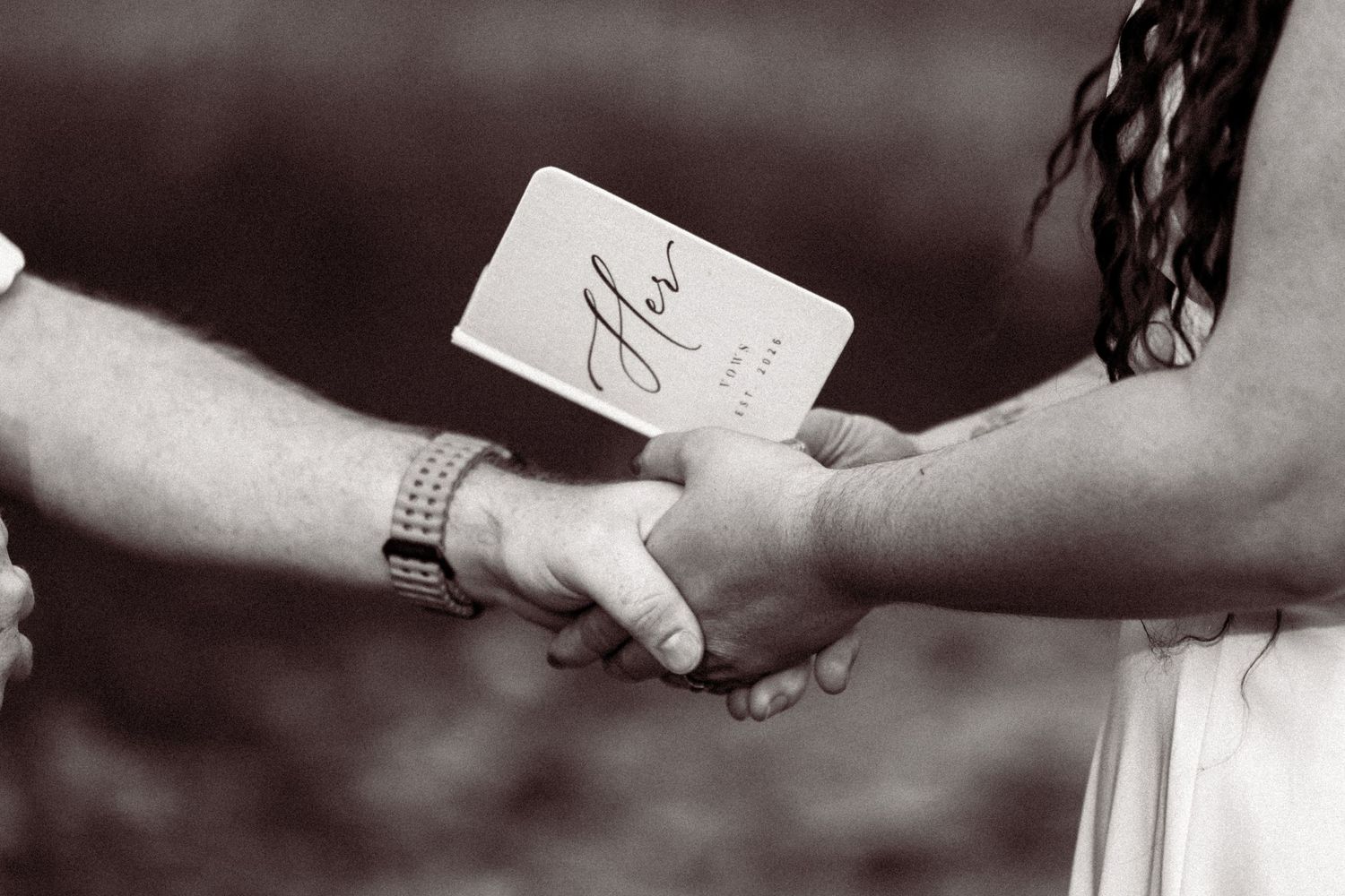 Black and white close-up of couple holding hands with wedding vow booklet during ceremony.