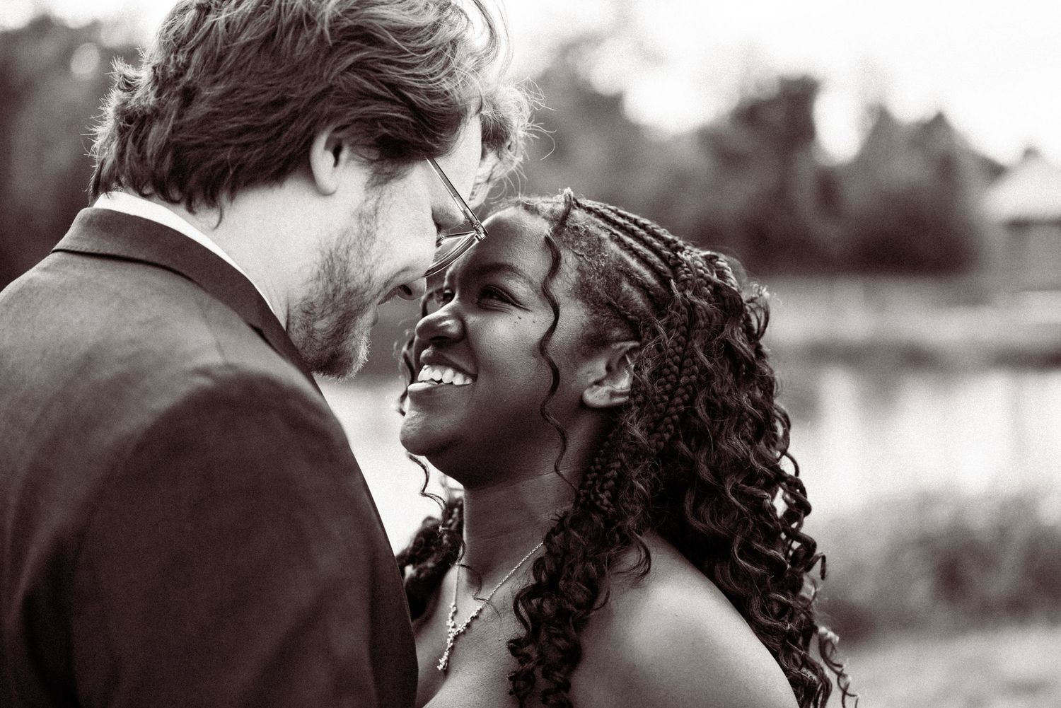 Black and white photo of an interracial couple sharing a romantic moment, smiling at each other outdoors.