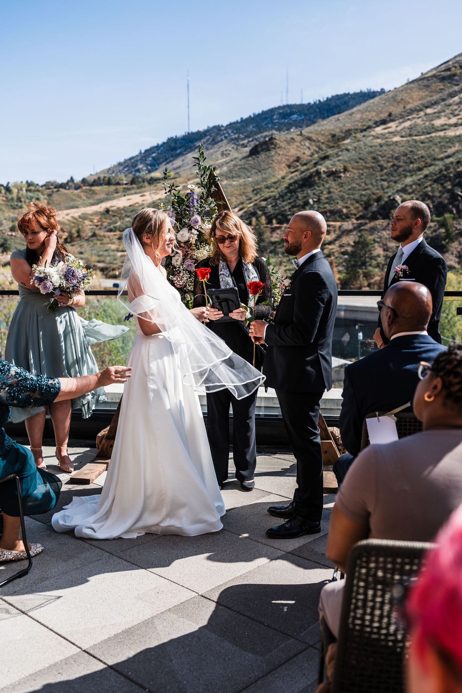 Outdoor wedding ceremony on a rooftop patio with mountain views, bride in white gown and groom in black suit.