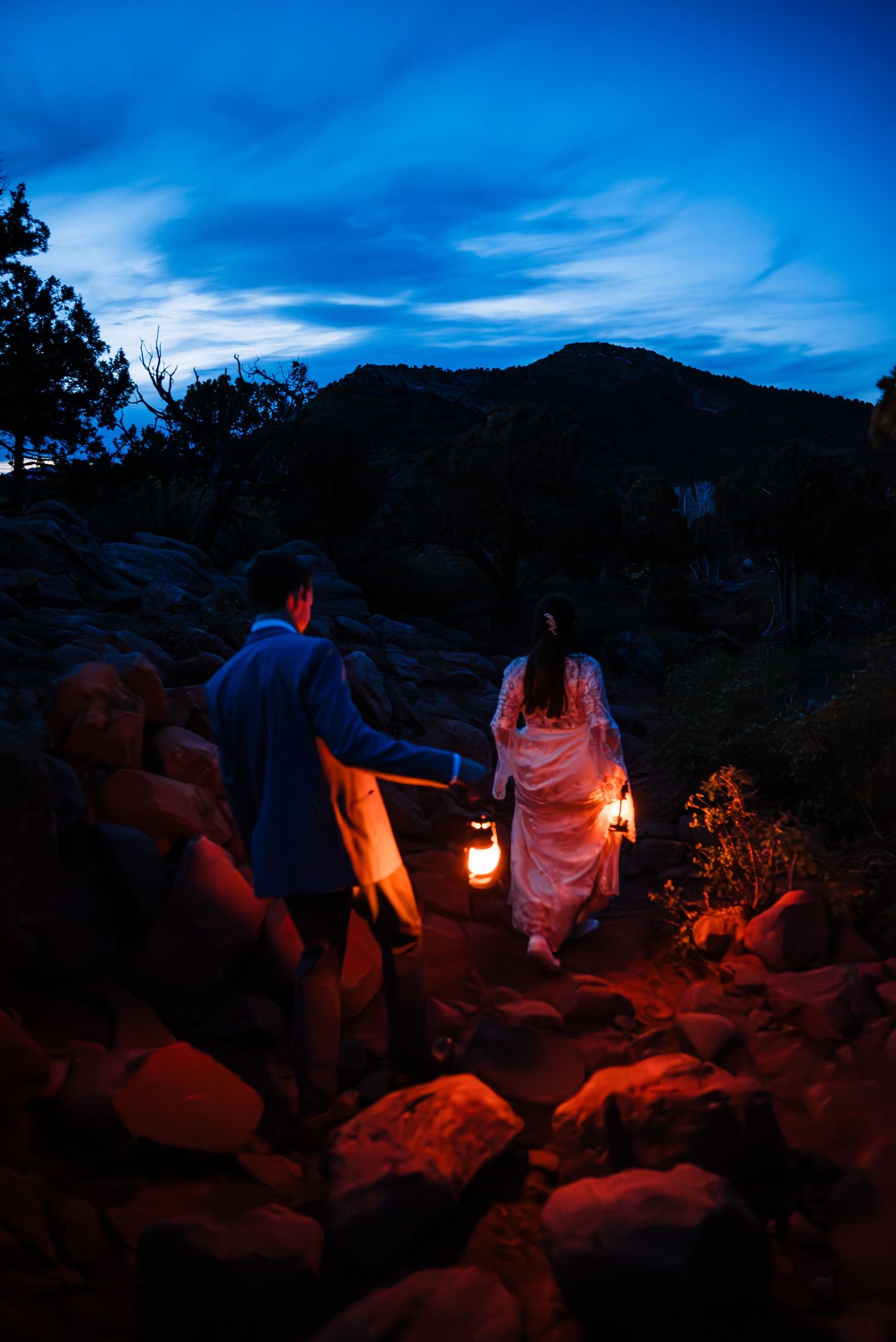 Bride and groom hiking rocky mountain trail at dusk, holding glowing lanterns in wedding attire.