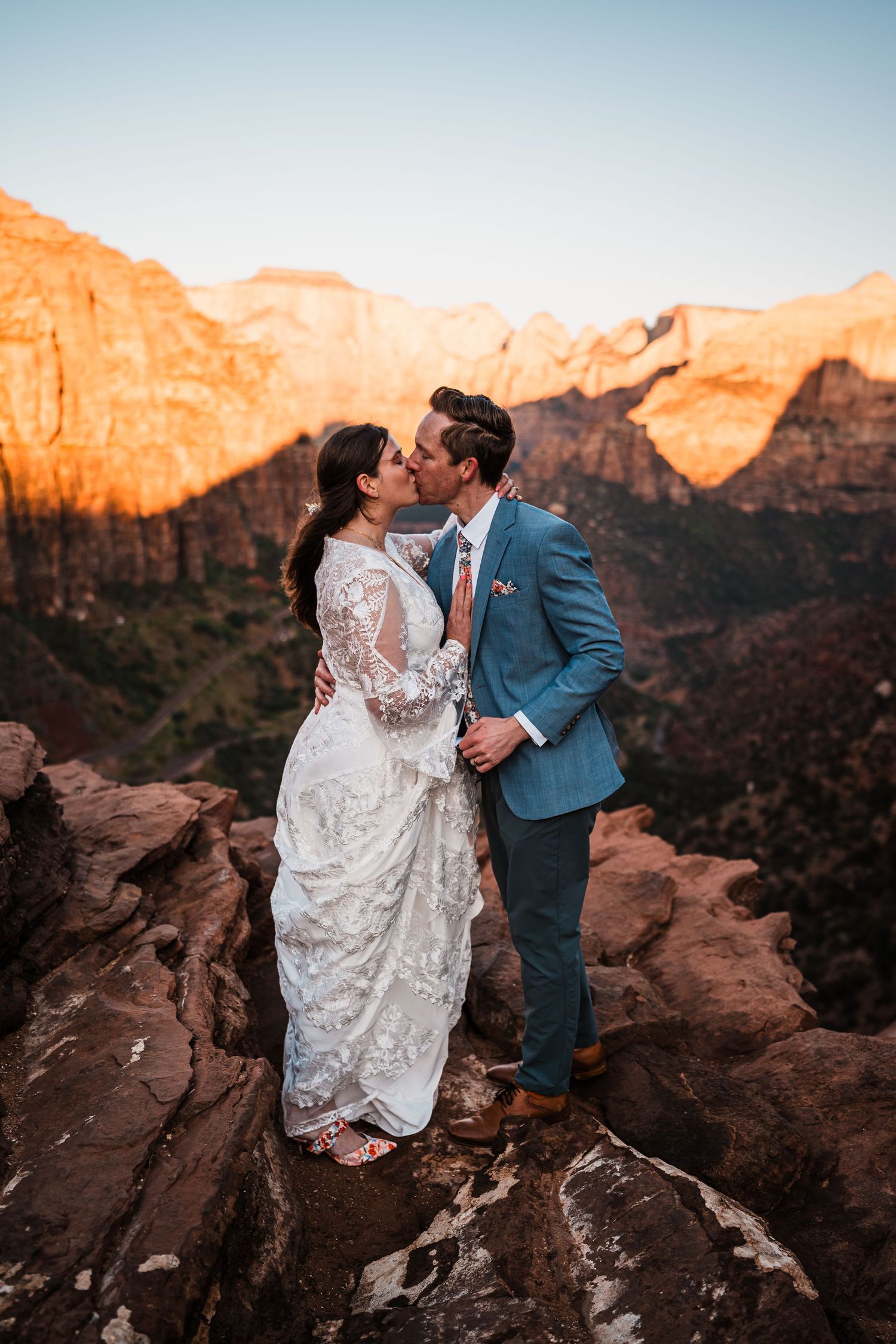 Newlyweds share a kiss on rocky cliff at sunset with stunning Zion National Park canyon views.