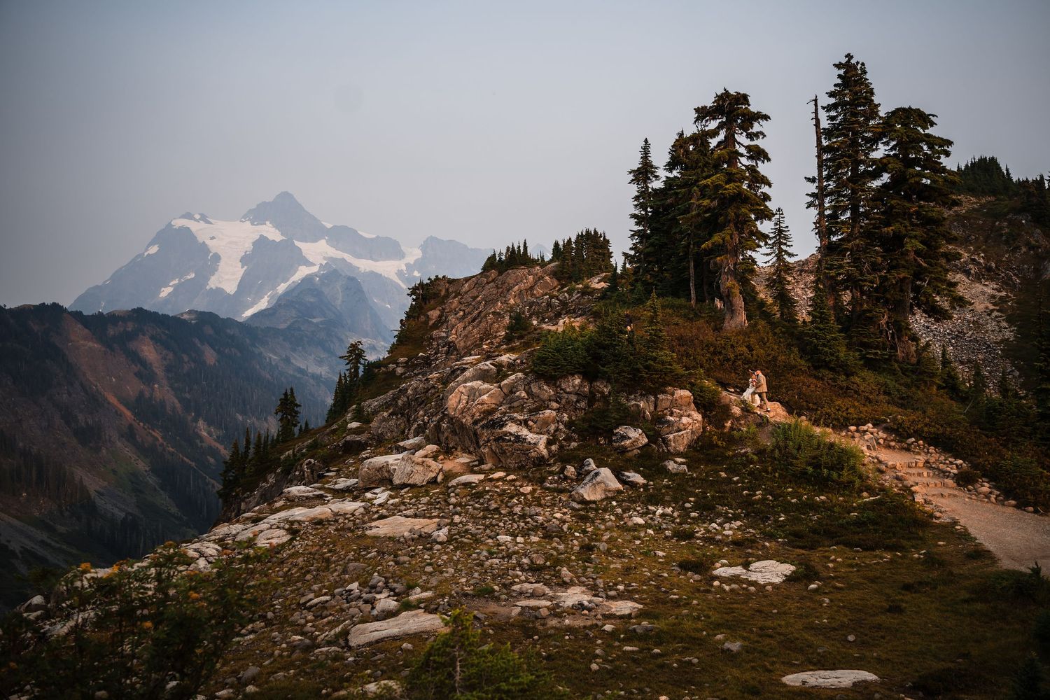 Scenic mountain landscape with snow-capped peaks, evergreen trees, and rocky terrain under cloudy skies.