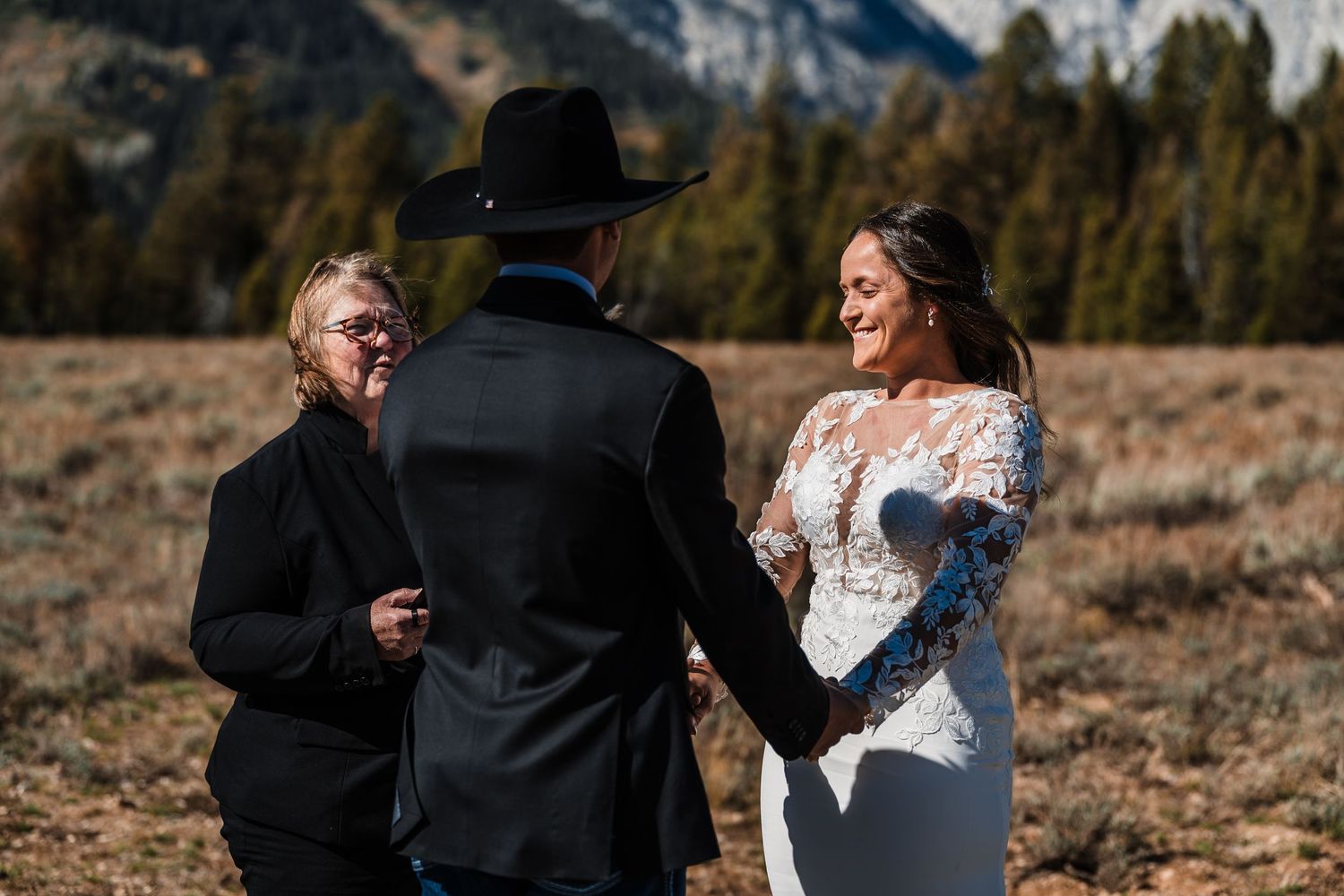 A couple exchanges vows during an outdoor wedding ceremony in a mountain meadow with evergreen trees in the background.