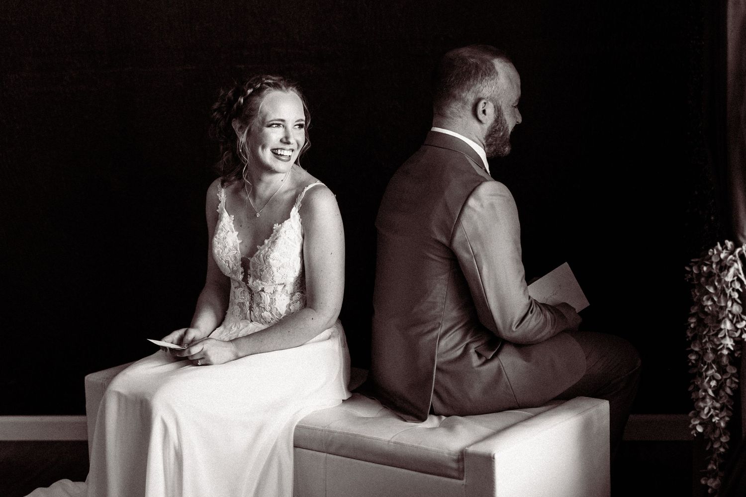 Black and white photo of bride and groom sitting back-to-back reading vows before ceremony.