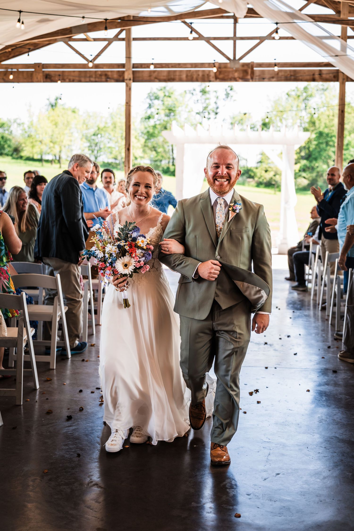 Smiling bride and groom walk down wedding aisle in rustic barn venue with colorful wildflower bouquet.