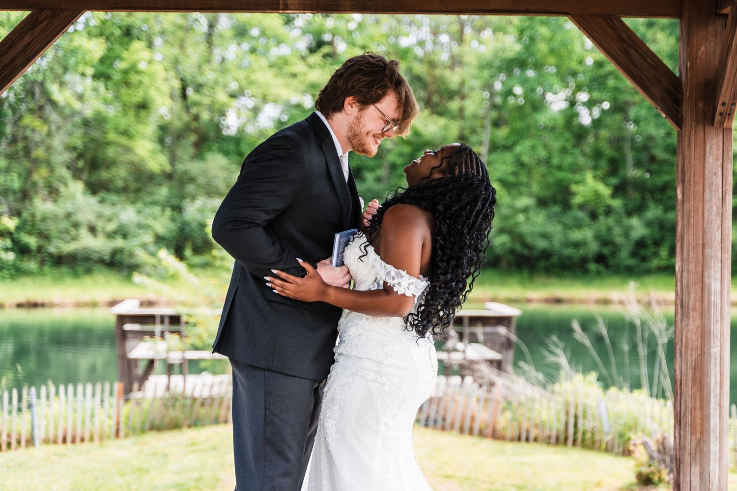 Interracial couple sharing a romantic embrace at an outdoor wedding venue near a pond.